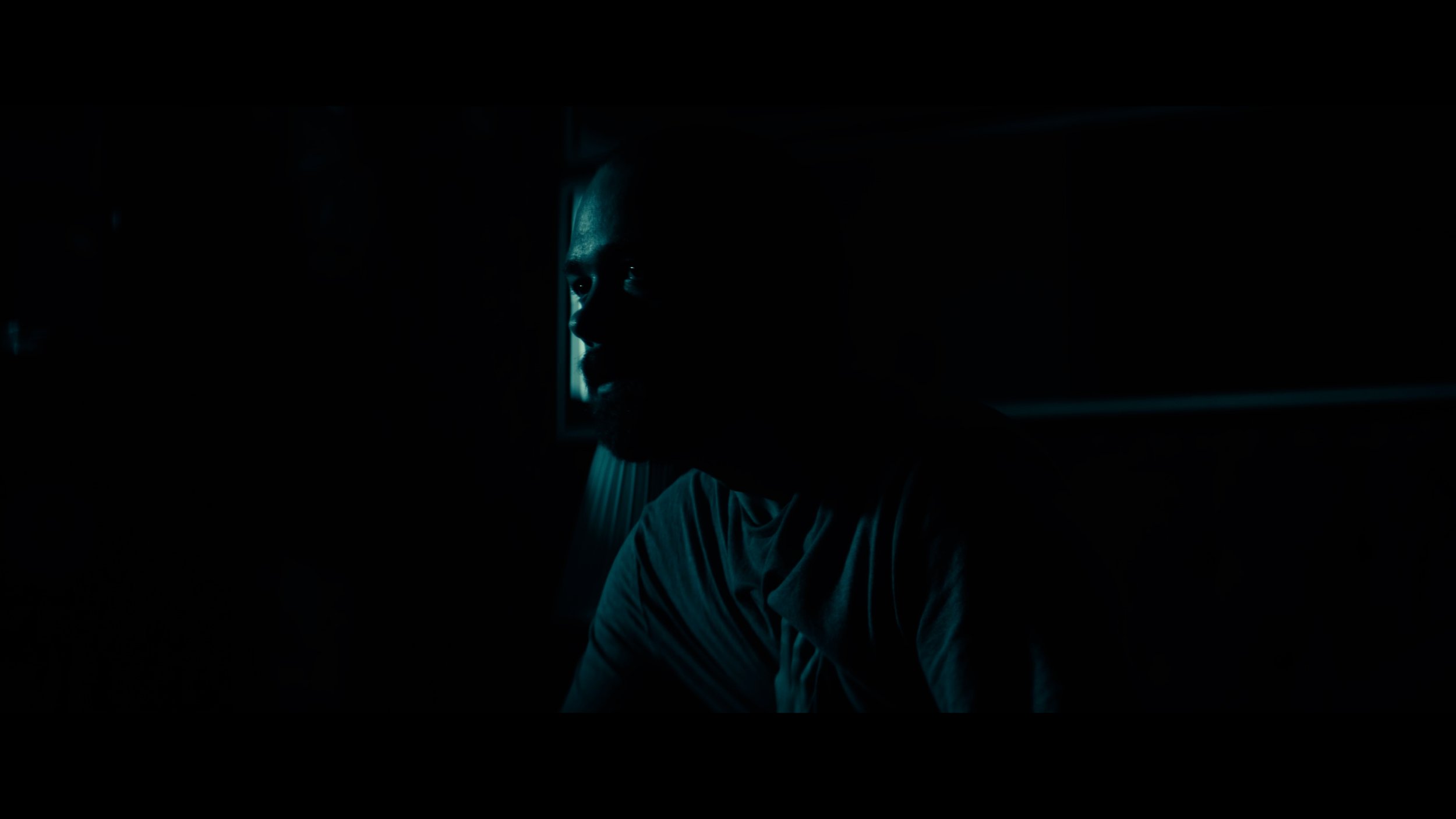 A person is partially visible in a dark room, with only part of their face illuminated by faint blue light, creating a moody and mysterious atmosphere.