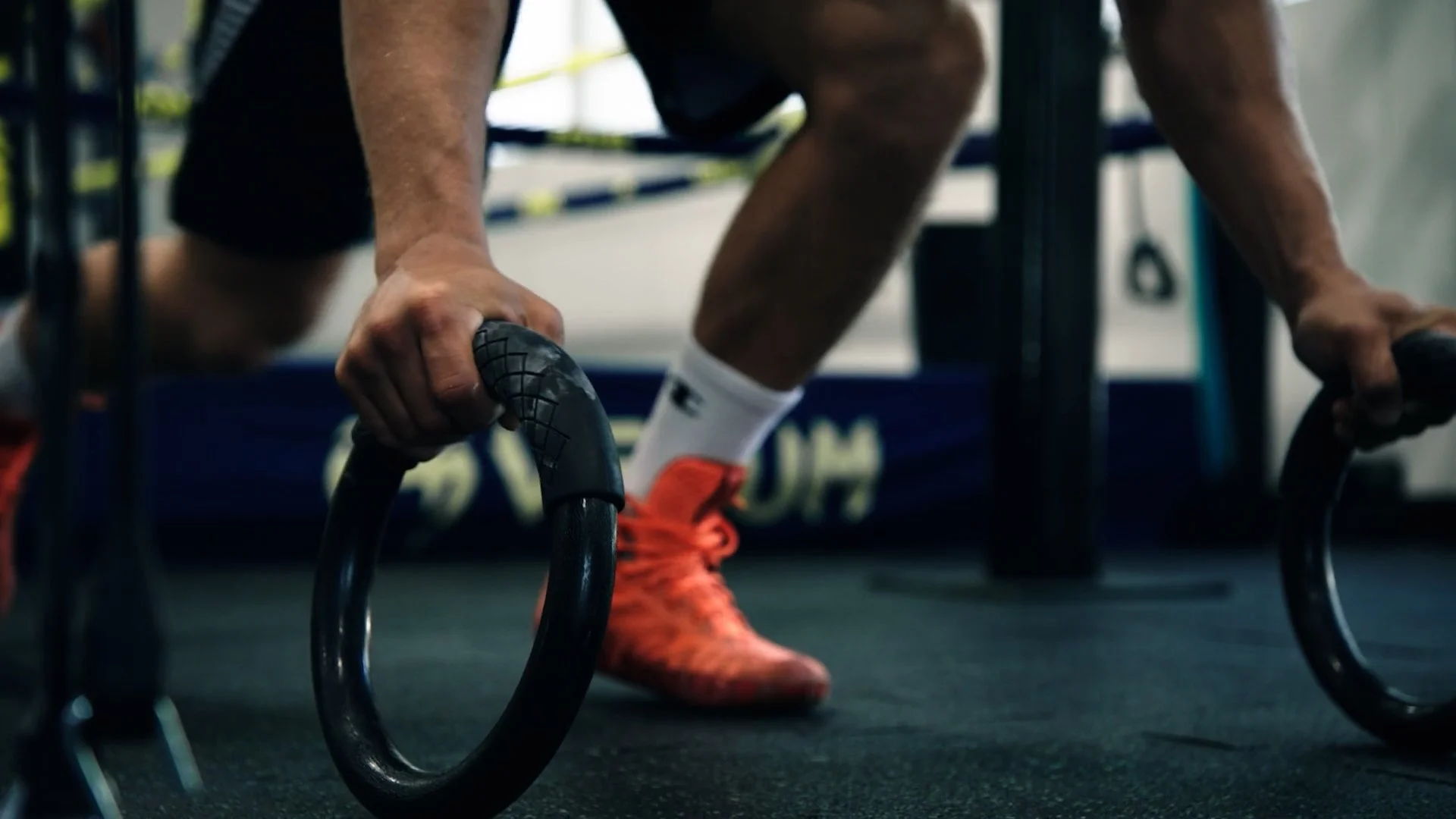 Close-up of a person gripping gymnastic rings, wearing orange sneakers and white socks, in a gym setting.