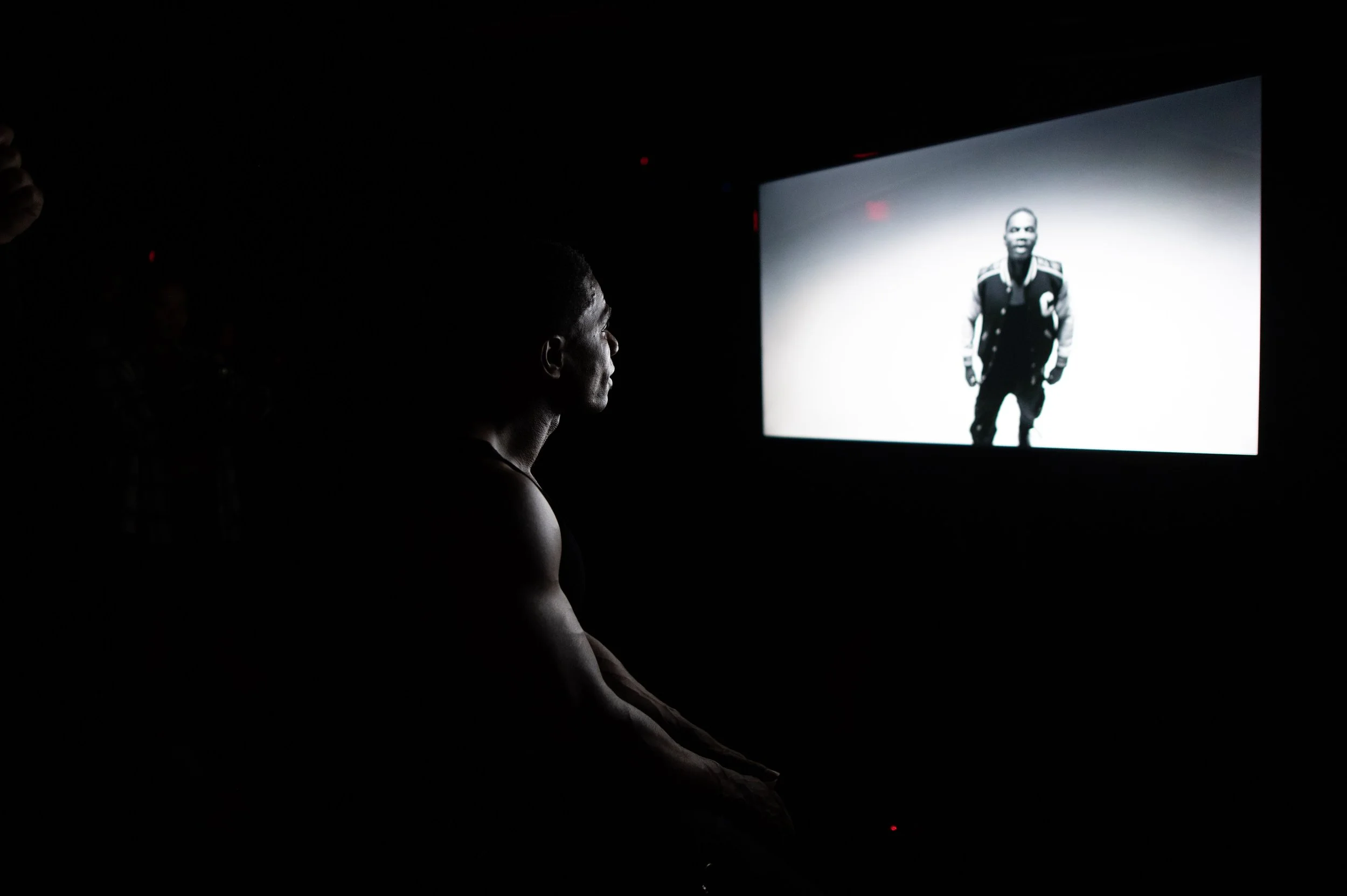 A person with a muscular build watches a black-and-white screen featuring a man in a varsity jacket.