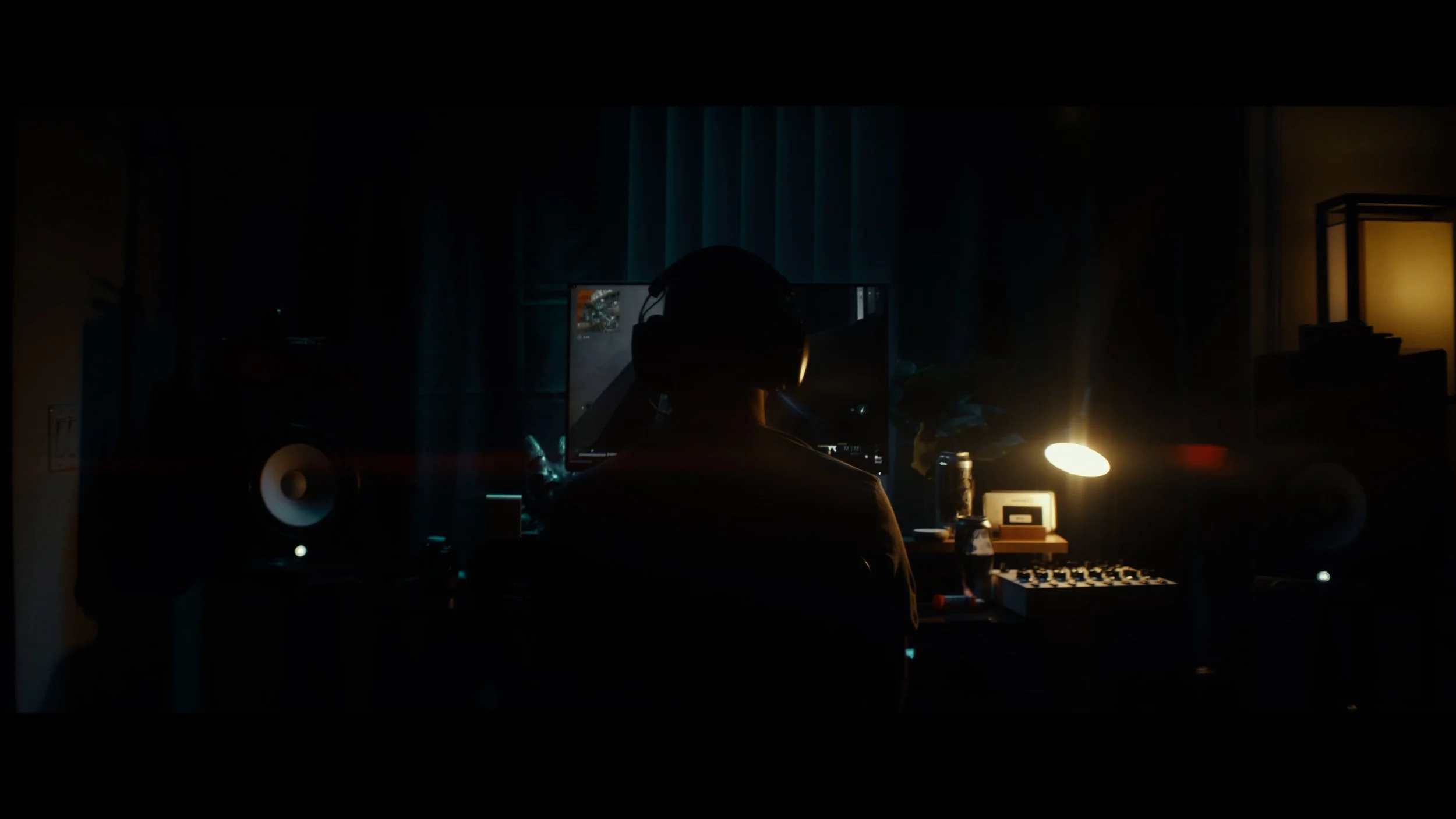 Person sitting at a desk with a computer monitor in a dimly lit room, wearing headphones, illuminated by a desk lamp, surrounded by audio equipment and supplies.