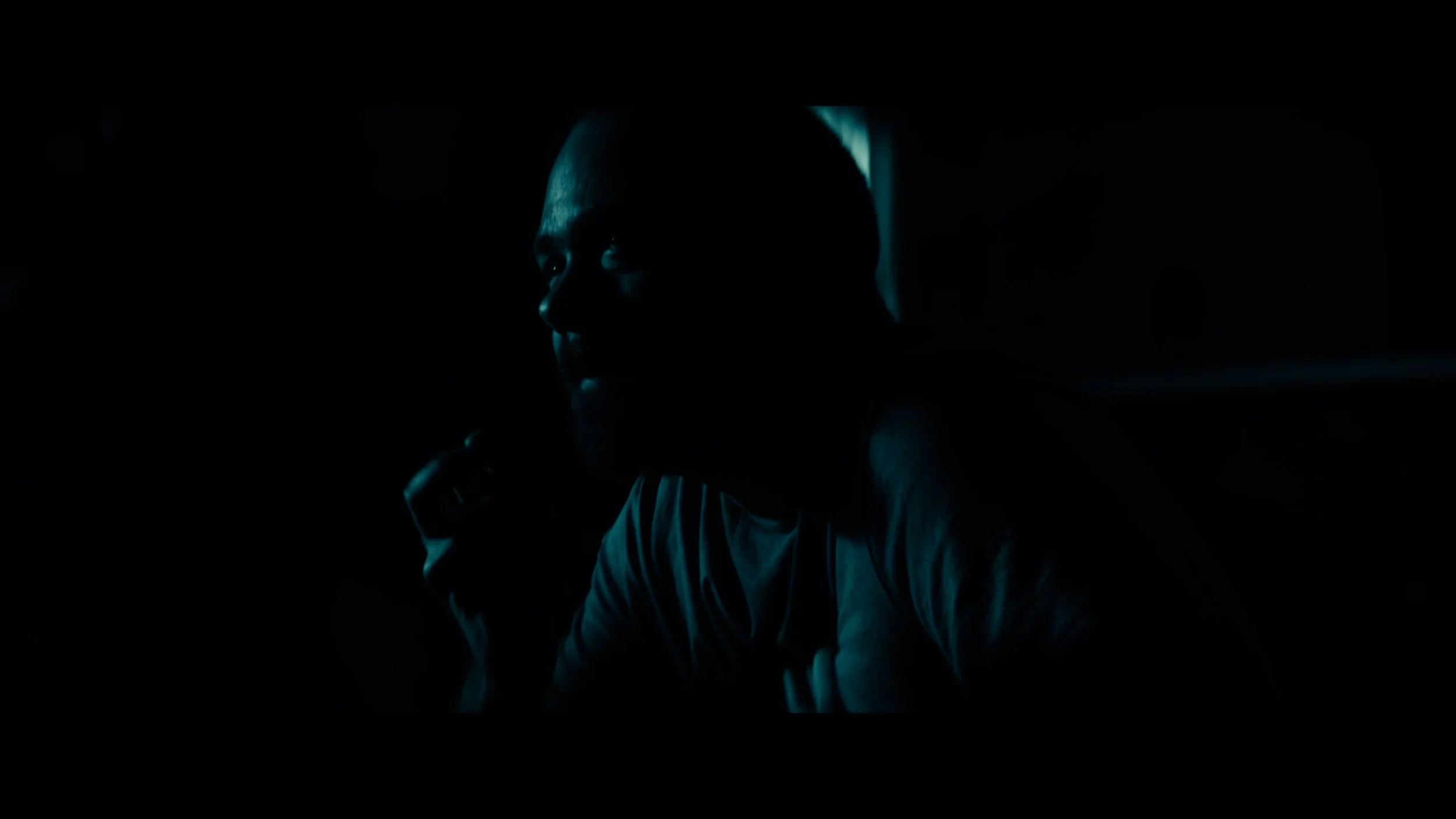 A close-up of a man in a dark room, illuminated by faint blue light, with a serious expression, holding an object near his mouth.