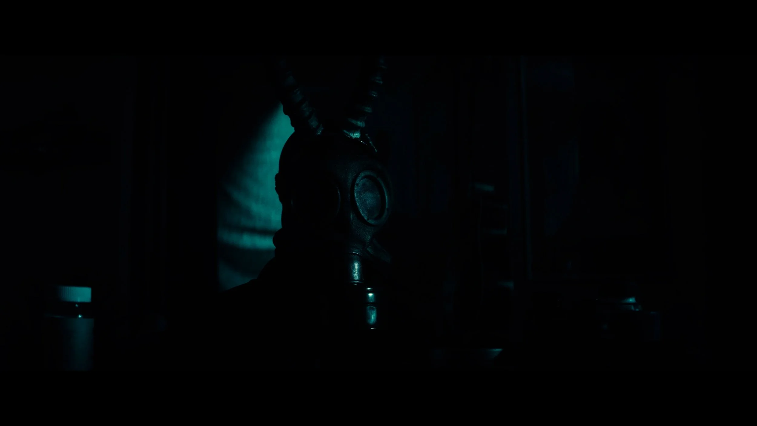 Person wearing a gas mask with horn-like protrusions, in a dark, shadowy environment with faint blue lighting.