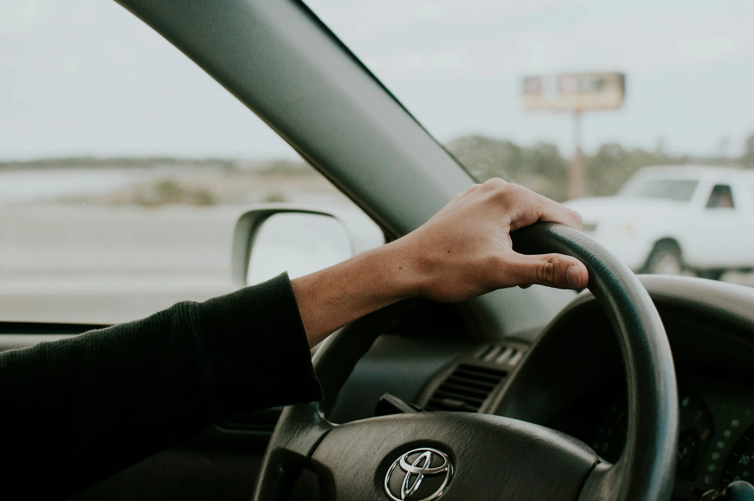 A person driving a Toyota vehicle, holding the steering wheel with one hand, with a view of a white vehicle on the road outside and a billboard in the distance.