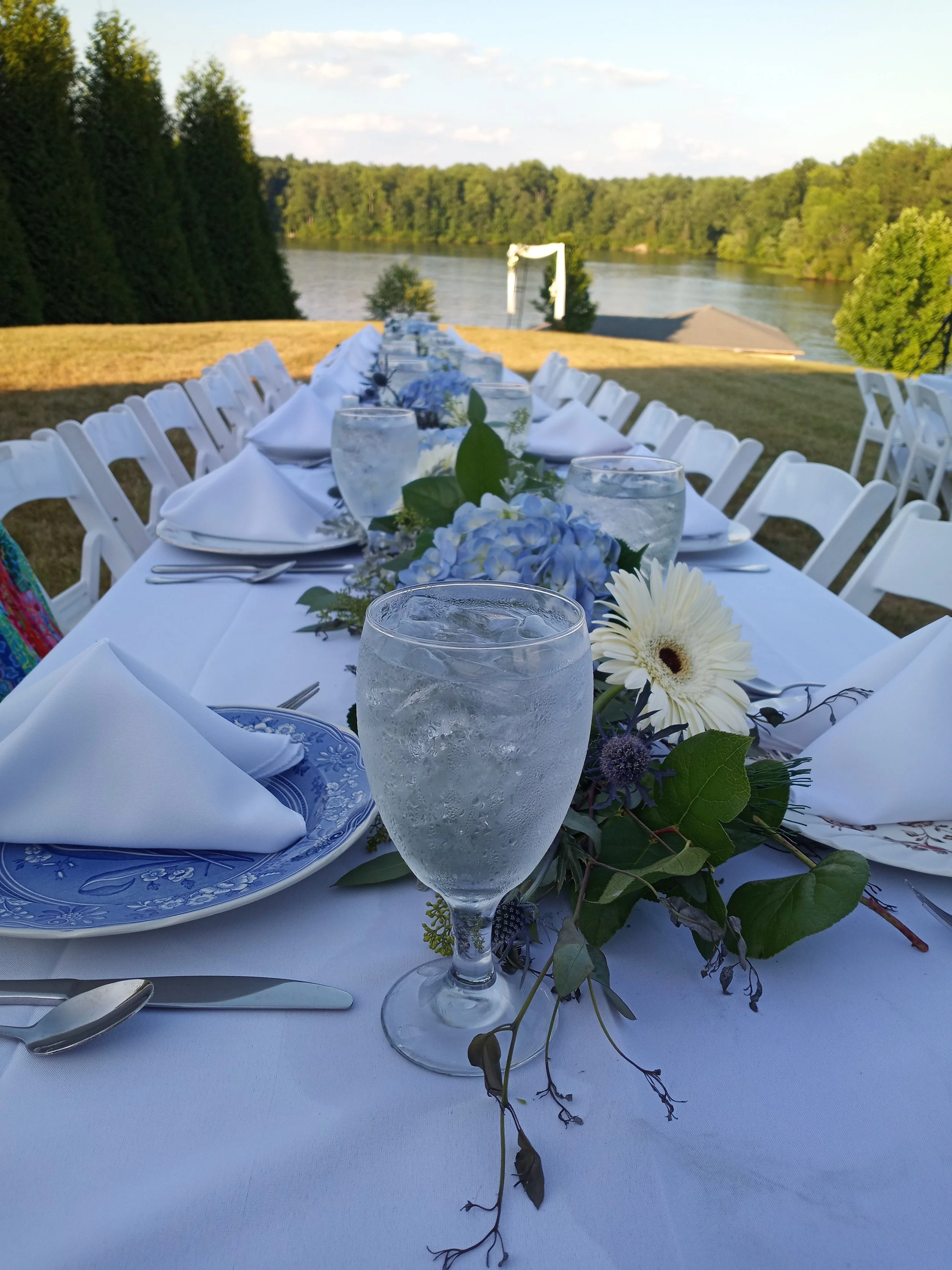 Elegant outdoor dinner table set up by a river with white chairs, floral centerpiece, water glasses, and white napkins