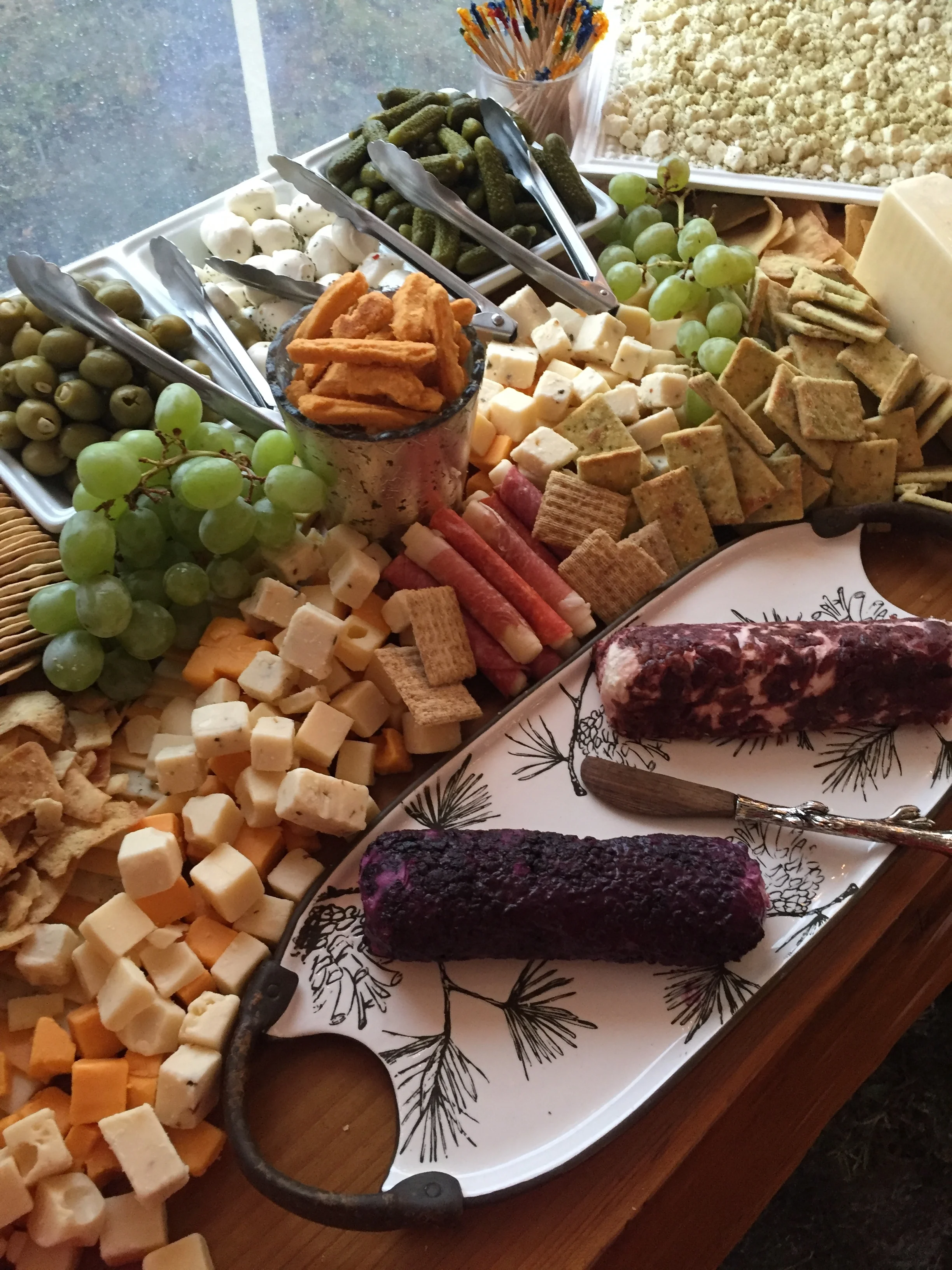 A cheese and charcuterie platter with grapes, cheeses, crackers, pickles, and dried sausage, with a small fork and toothpicks.
