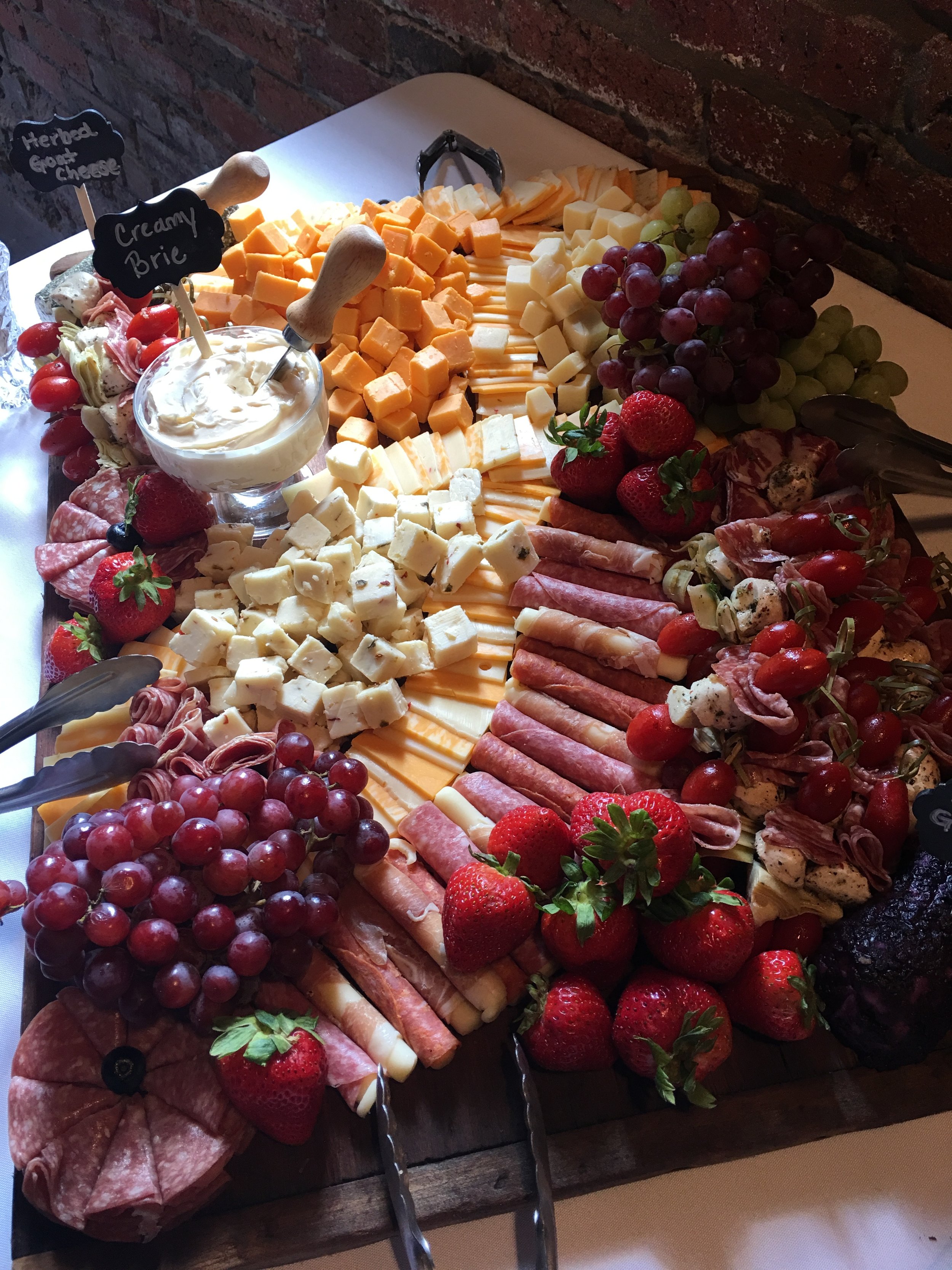 Cheese and meat platter with grapes, strawberries, and a bowl of herb and goat cheese spread, with small chalkboard signs labeling herbs and creamy brie.