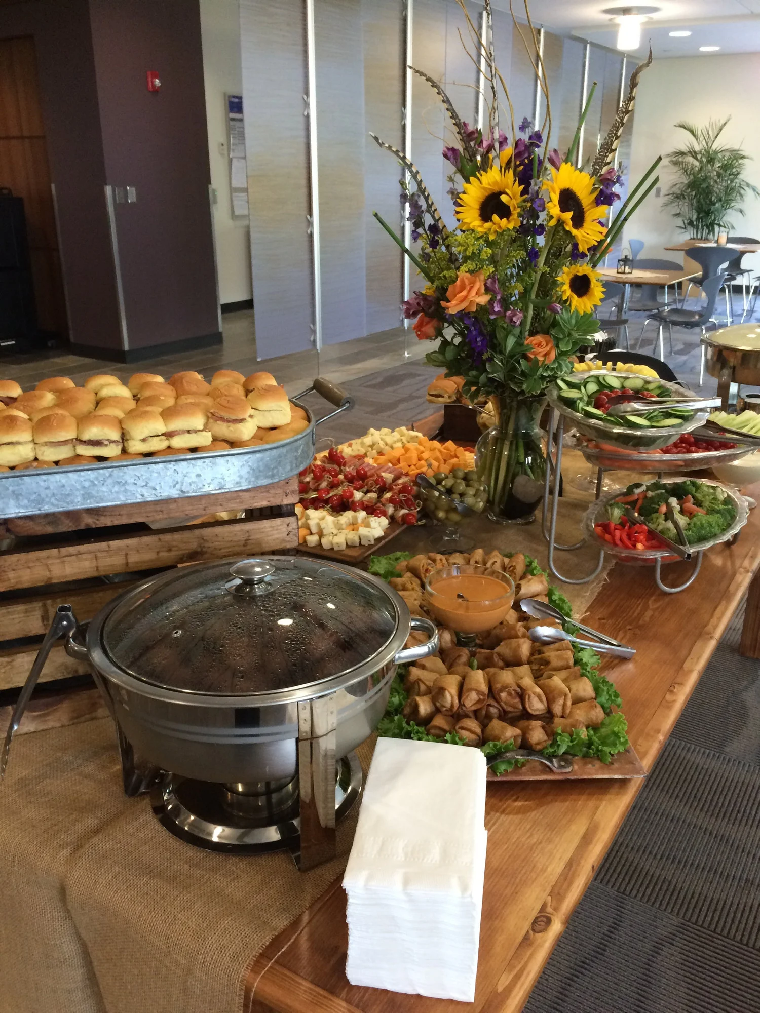 Buffet table with mini sandwiches, spring rolls, salads, grapes, cheese, cherry tomatoes, cucumber slices, and a large bouquet of sunflowers and purple flowers.