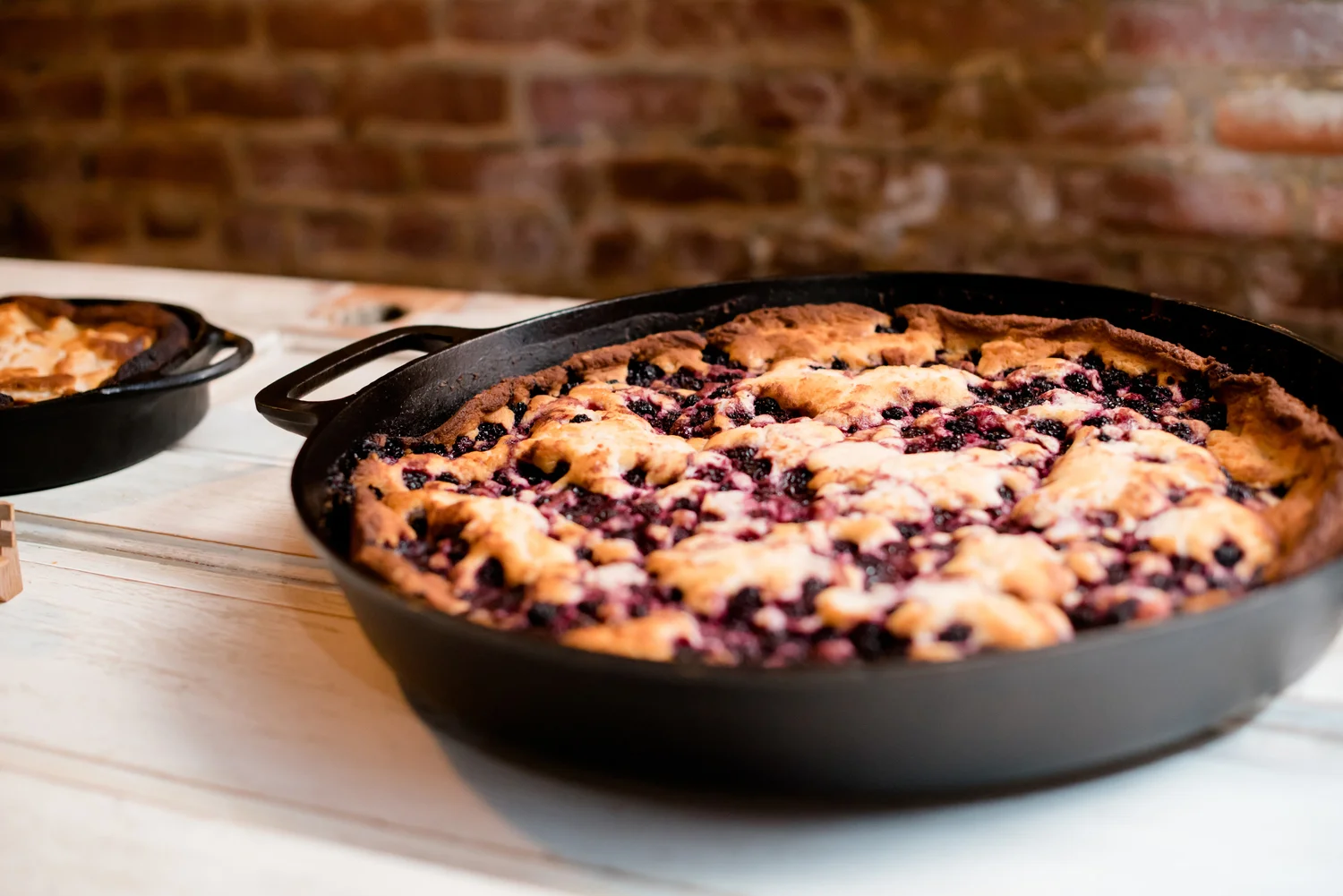 Cast-iron skillet containing a freshly baked blueberry cobbler with a golden crust, brick wall in the background.