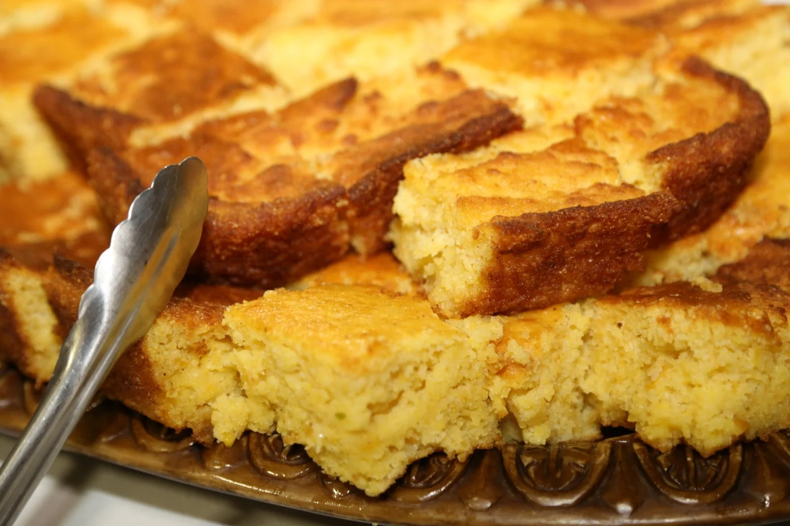 A close-up of a sliced piece of cornbread or similar baked dish on a decorative plate with a pair of tongs.