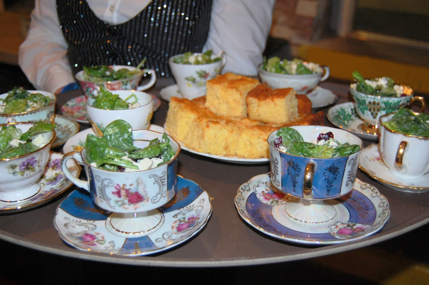 Tea set with floral china cups and saucers filled with green salad, and a plate of cornbread or cake slices on a tray.