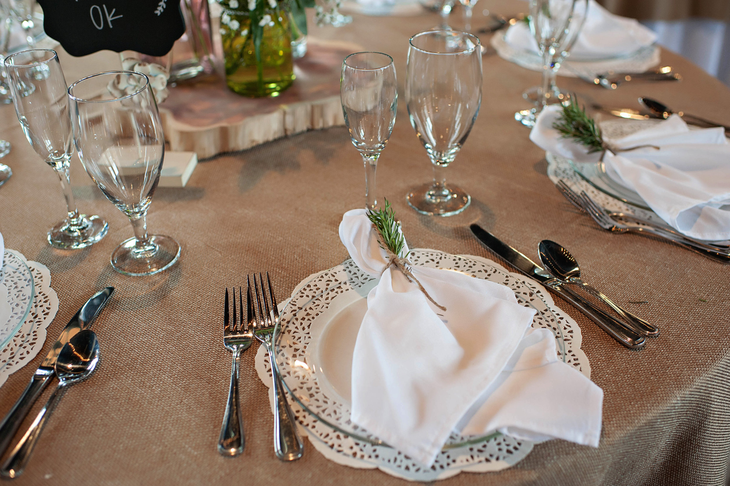 A formally set dining table with white ceramic plates, silver cutlery, clear wine glasses, a beige tablecloth, and white napkins decorated with small green sprigs, possibly rosemary. There is a wooden centerpiece with a small black chalkboard sign that has the word 'OK' written on it.