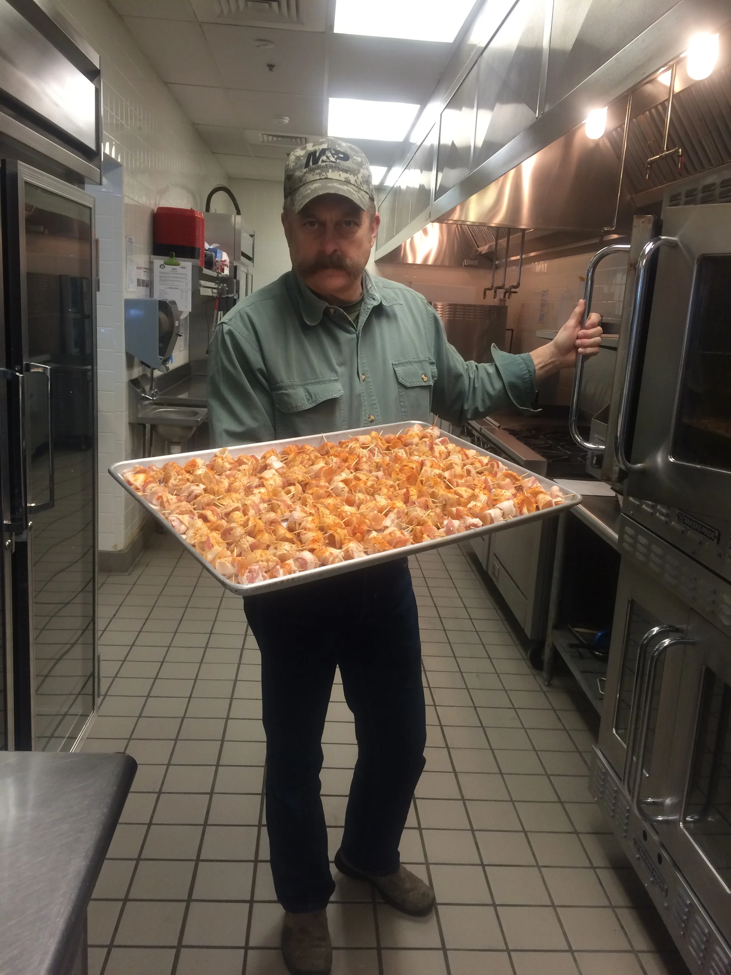 A man wearing a camouflage cap and green shirt holding a tray of raw chicken pieces in a commercial kitchen.