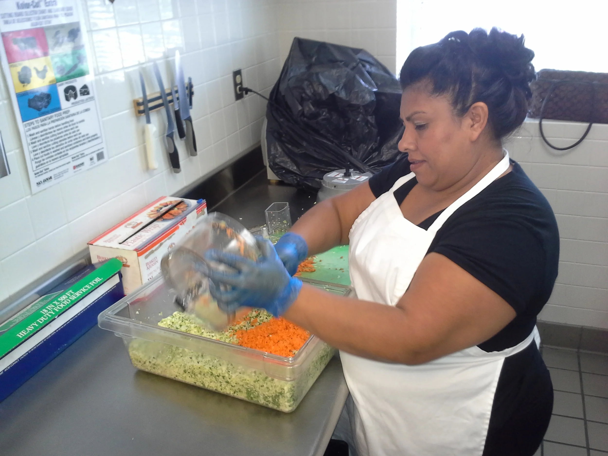 Woman wearing a white apron and blue gloves preparing vegetables in a commercial kitchen.