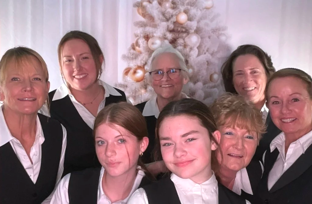 Nine women, including two young girls, posing together in front of a white Christmas tree decorated with gold and white ornaments, celebrating during the holiday season.