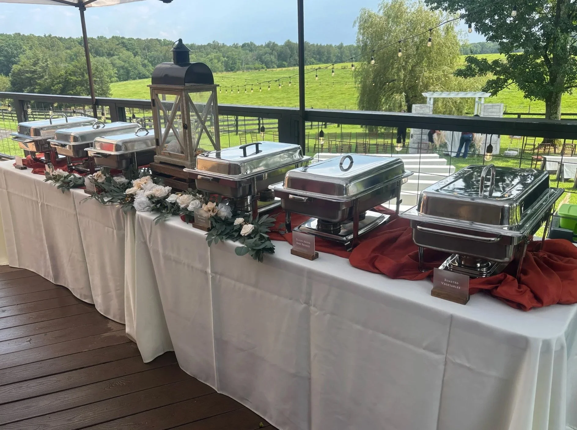 Buffet table with chafing dishes labeled for food items, decorated with white flowers and greenery, set outdoors on a wooden deck with scenic green landscape and string lights in the background.