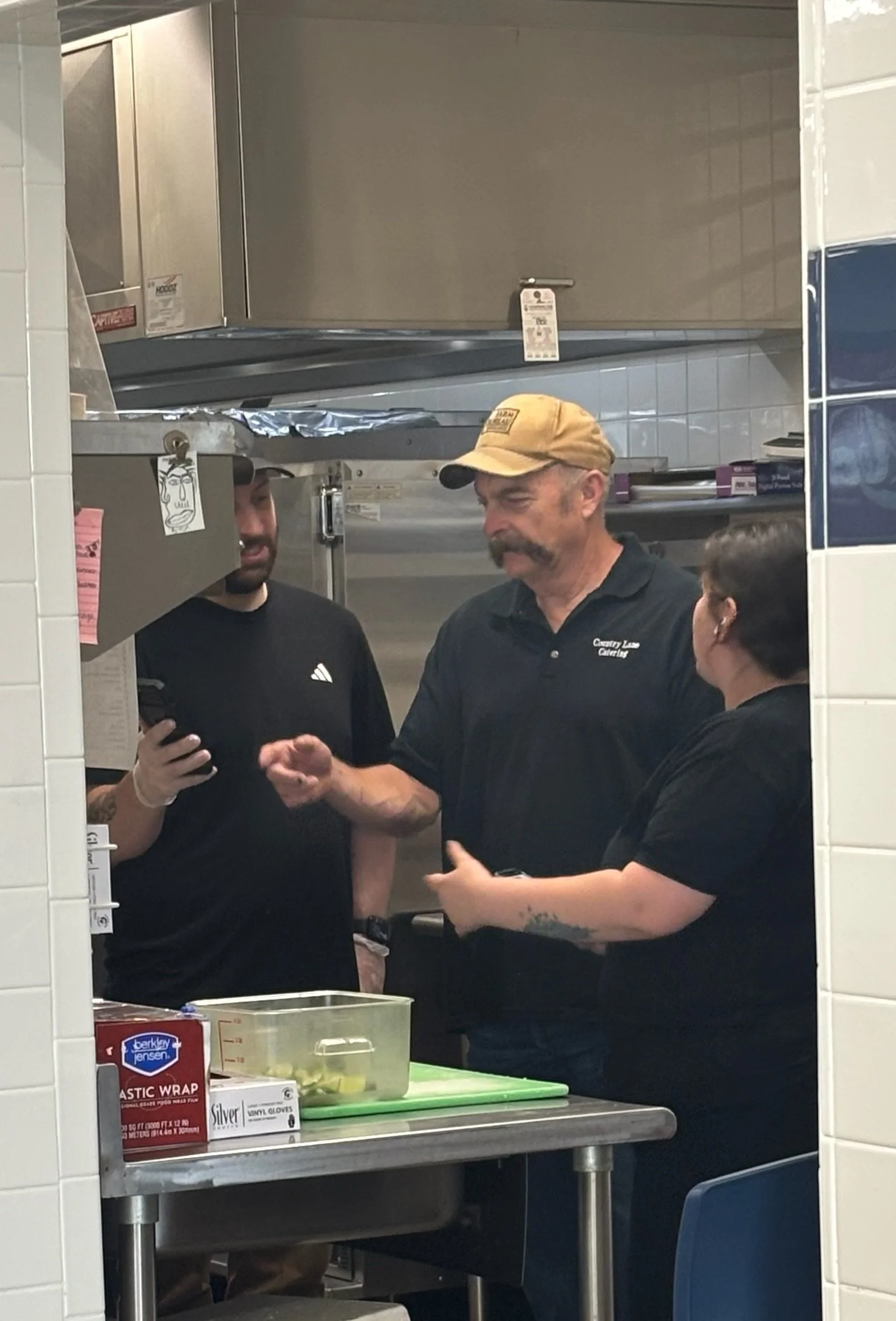 Three people, two men and one woman, having a conversation in a kitchen or food preparation area. One man is wearing a tan baseball cap, a black polo shirt, and has a mustache. The woman has dark hair, glasses, and a tattoo on her arm. The man in the black t-shirt has a beard and is holding a phone.