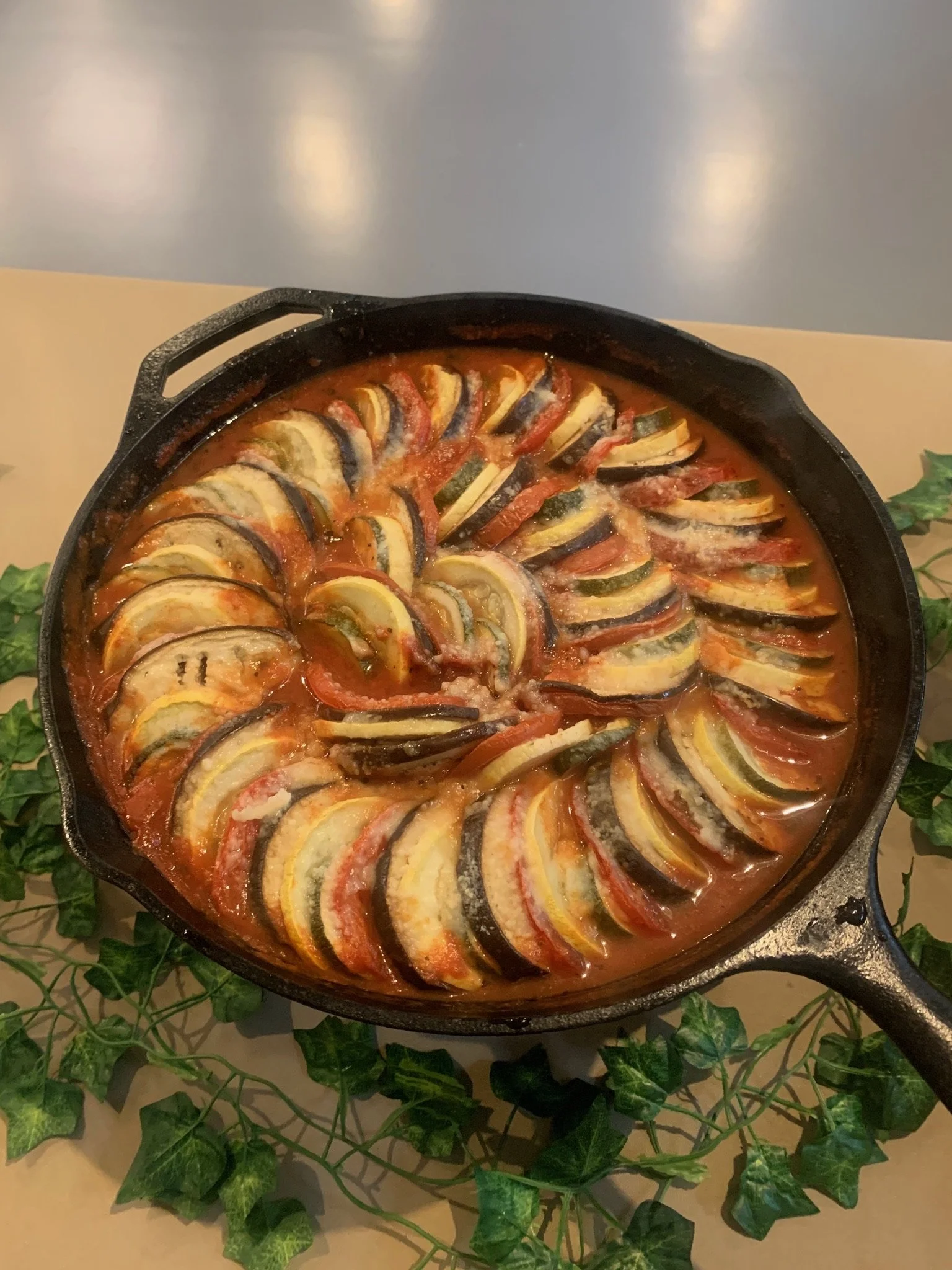 A cast iron skillet containing a layered vegetable and tomato dish, arranged in a circular pattern, on a table with green ivy decorations.