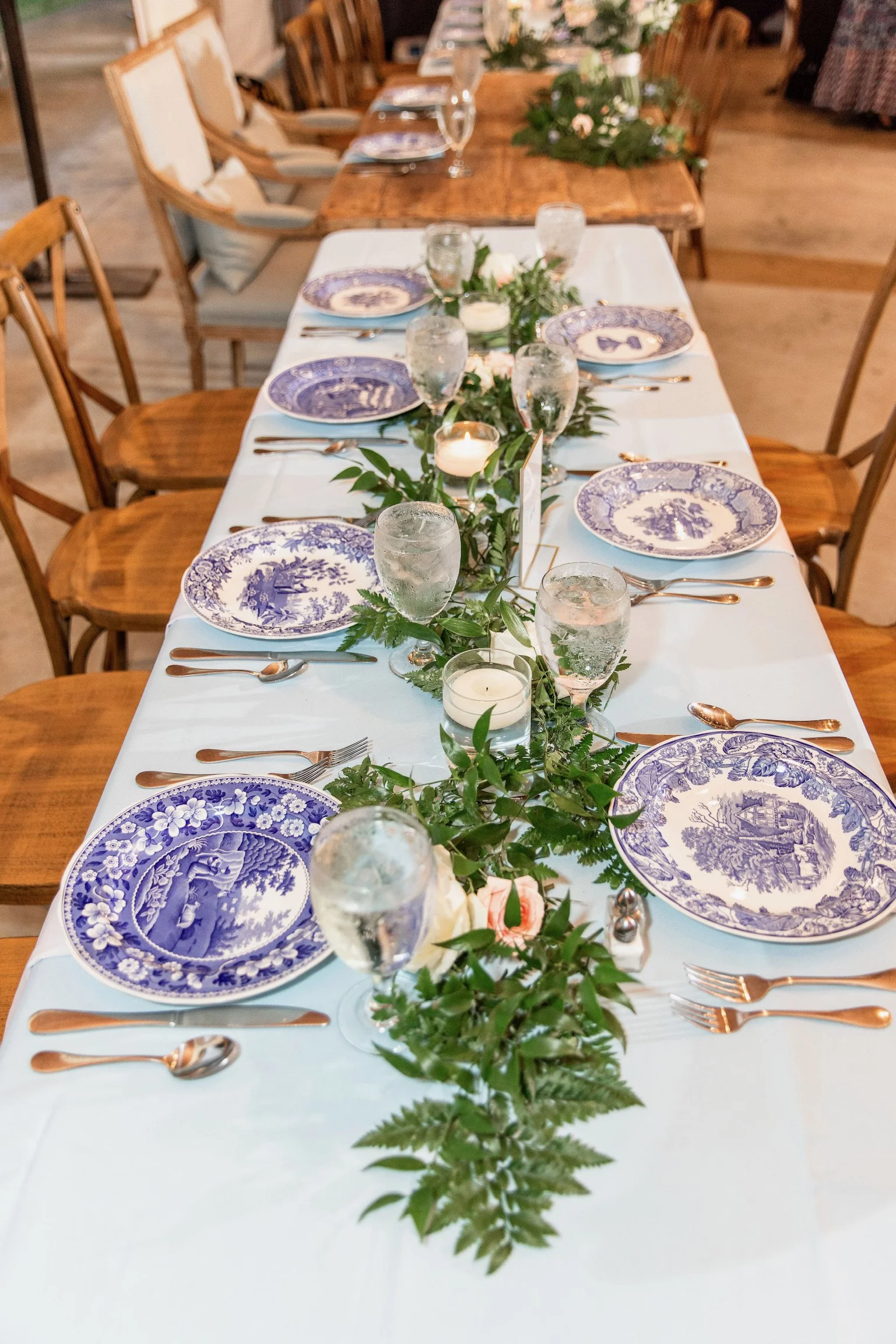 Wedding or formal event dining table set with blue and white patterned plates, silverware, water glasses, white candles, and green foliage garland.
