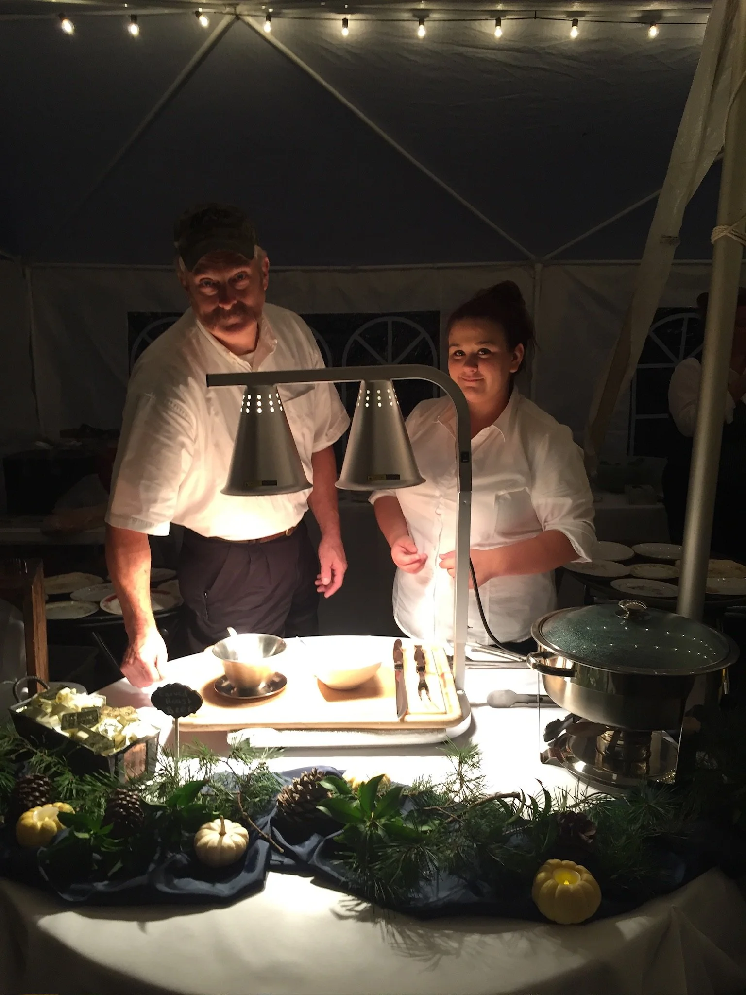 Two chefs standing behind a table with a heating lamp, preparing food at a dinner event inside a tent decorated with string lights and foliage.