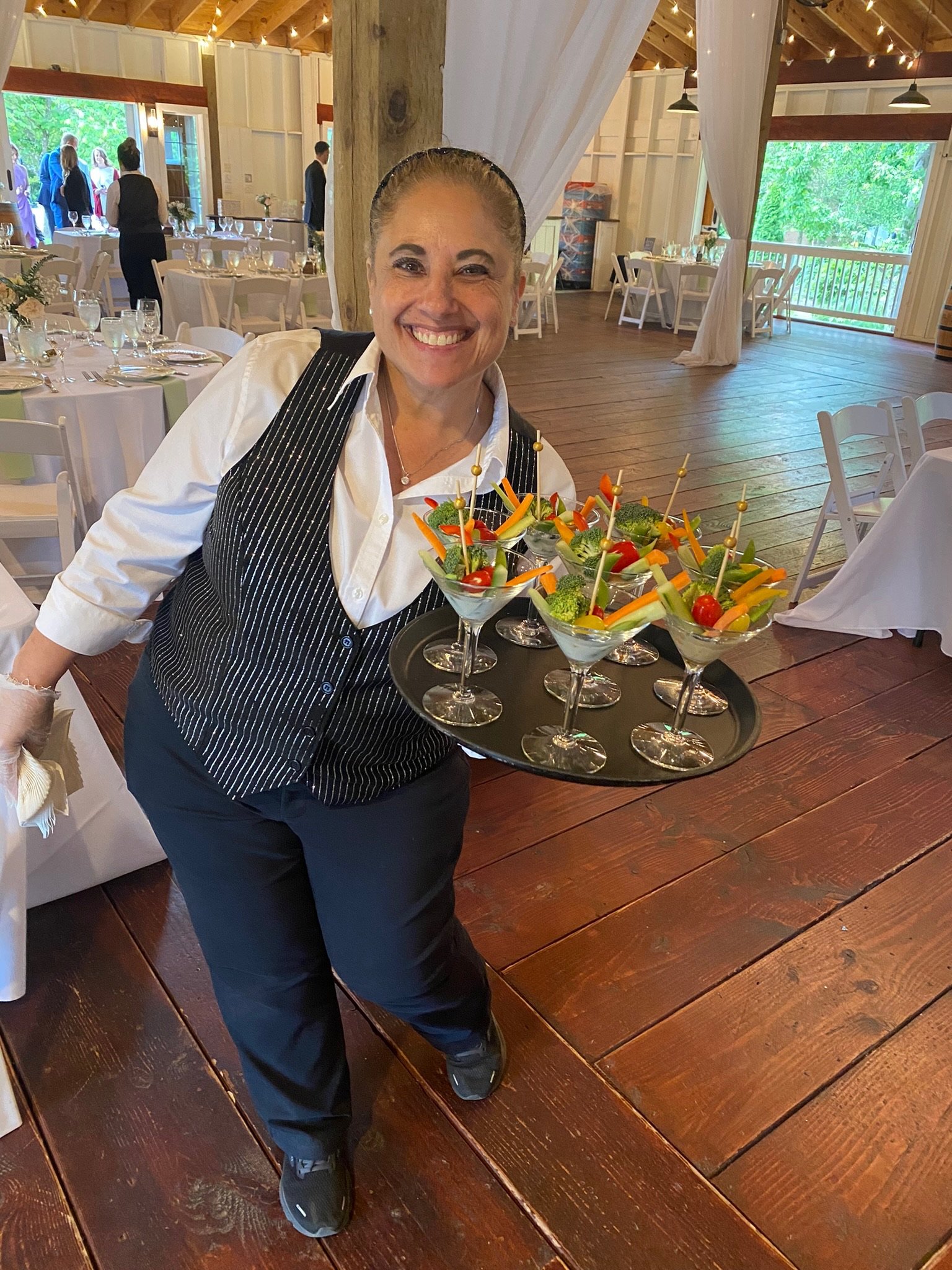 A smiling woman in a white shirt, black striped vest, and black pants holding a tray of colorful vegetable snacks in an event hall with tables, chairs, and other guests in the background.