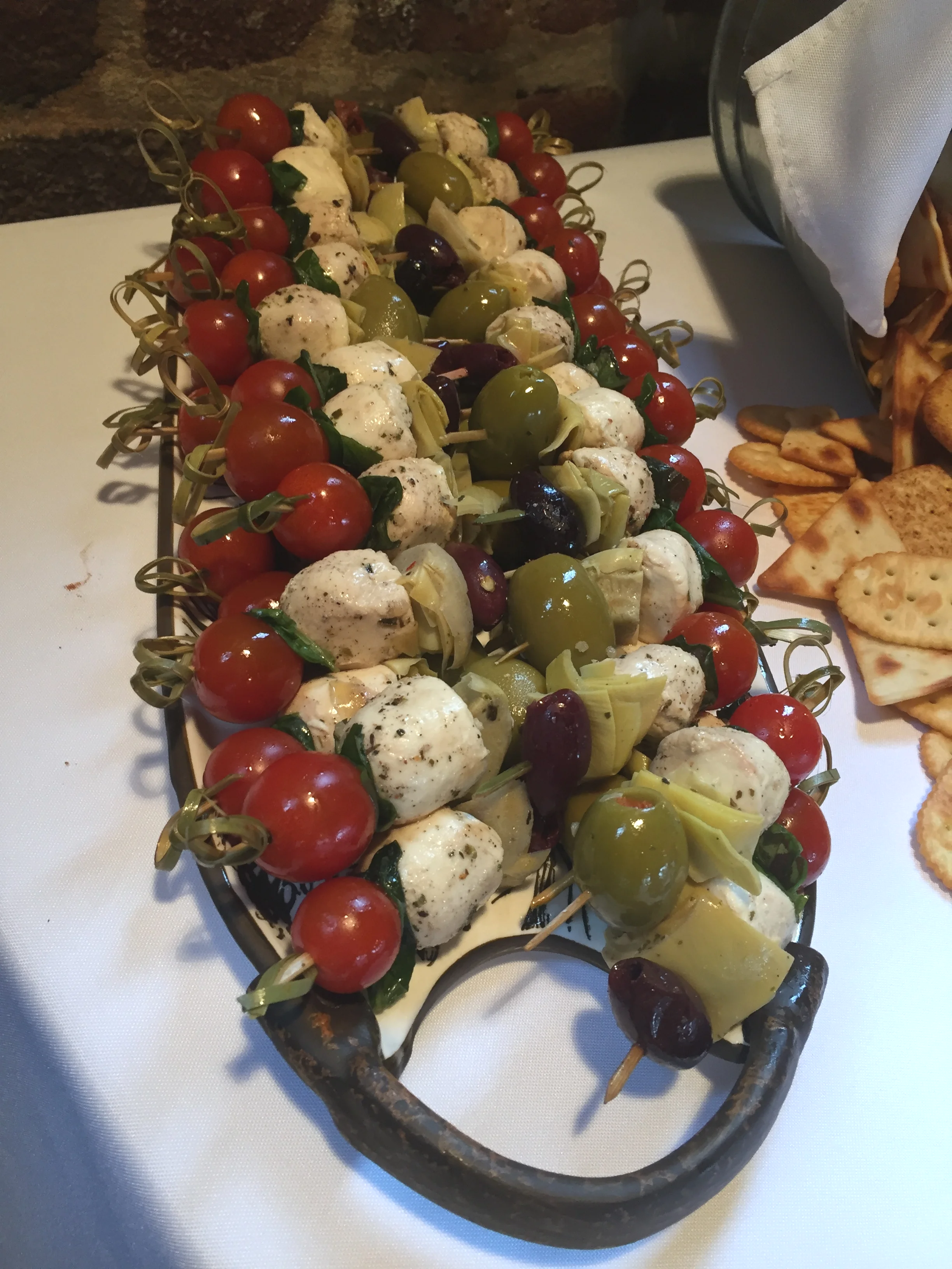A serving platter of assorted Caprese skewers with cherry tomatoes, mozzarella balls, green olives, black olives, artichoke hearts, and basil leaves.