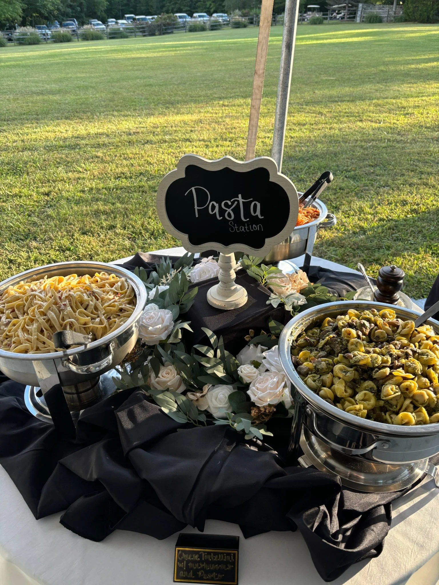 Contemporary pasta station with two large serving bowls of pasta, one with fettuccine and the other with a mixture of pasta and vegetables, decorated with white roses and greenery, set outdoors on a table with black and white fabrics, and a small chalkboard sign reading 'Pasta Station' in the background.