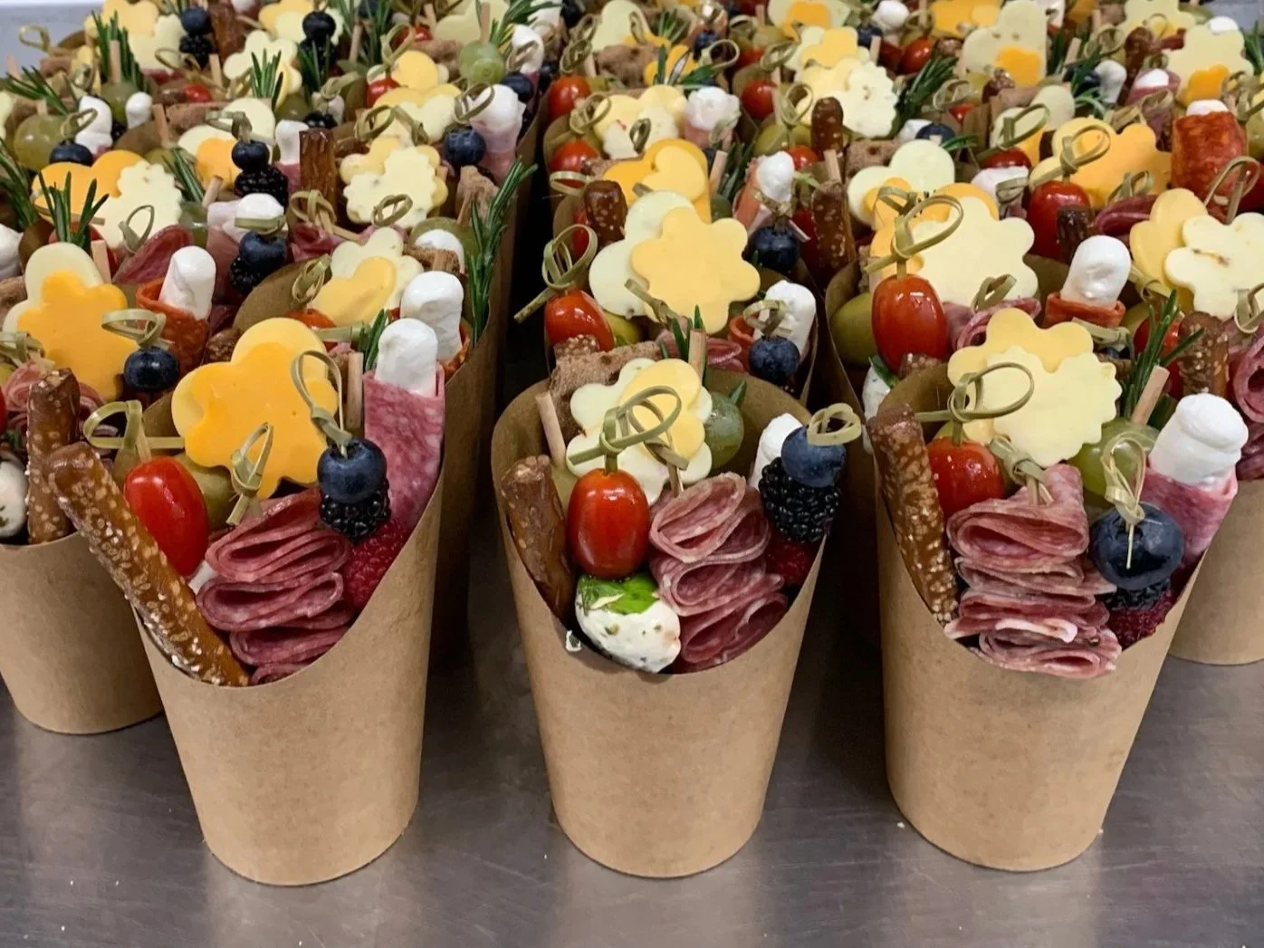 Multiple small paper cones filled with assorted charcuterie items including meats, cheese, grapes, cherry tomatoes, blackberries, and olives, arranged in a neat display.