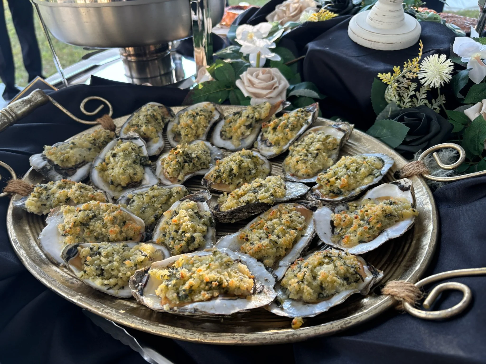 A silver tray of baked oysters topped with breadcrumbs and herbs on a decorative black and gold tablecloth with floral arrangements in the background.