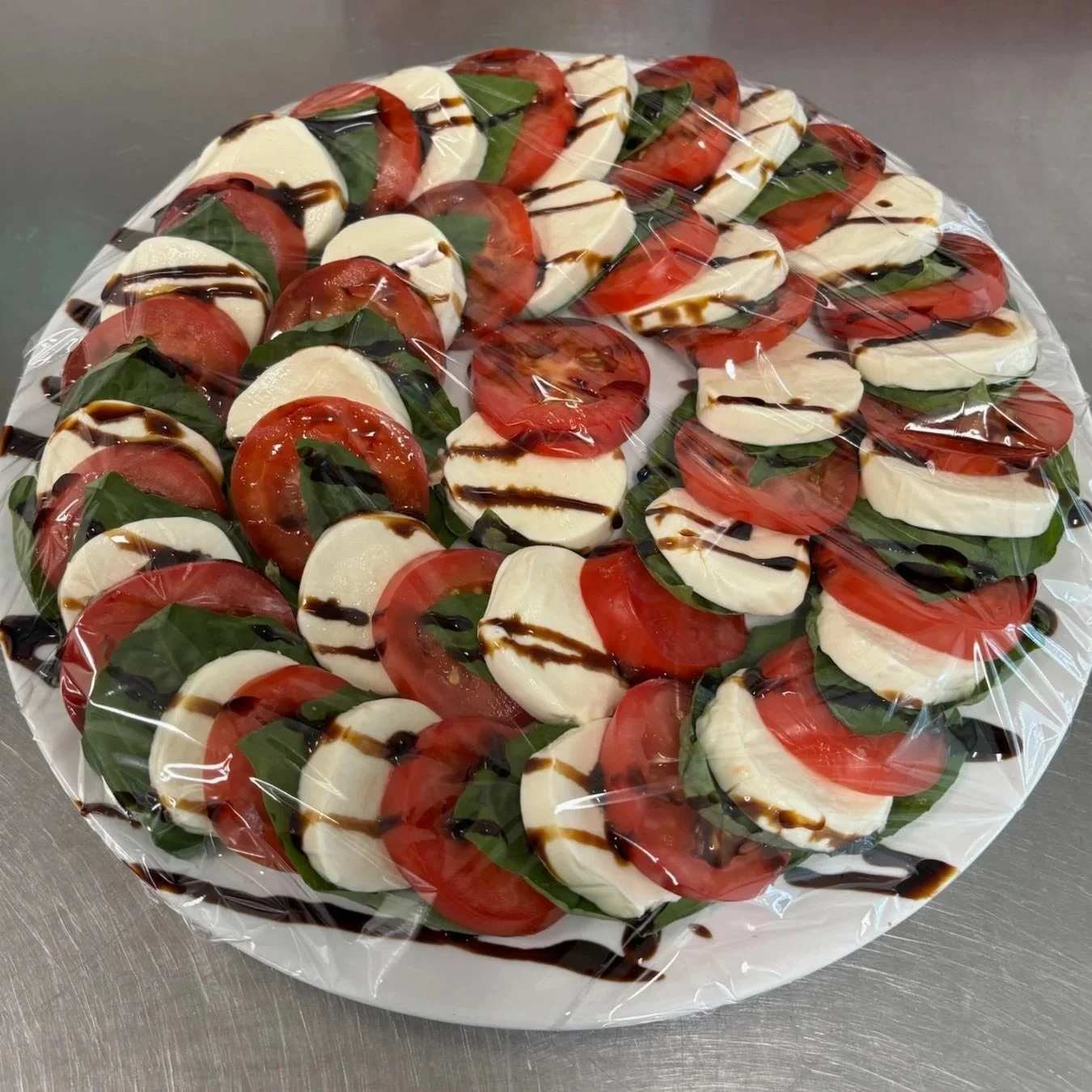 Caprese salad with sliced tomatoes, fresh mozzarella, basil, and balsamic glaze on a plate covered with plastic wrap.