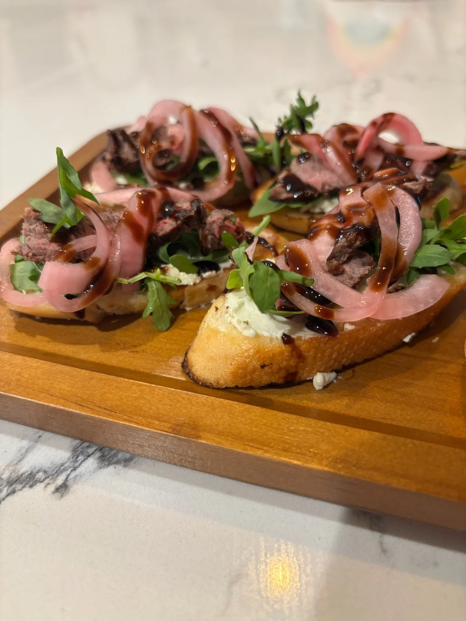 Open-faced sandwiches topped with beef, pickled onions, greens, and drizzle of balsamic glaze on wooden serving board.