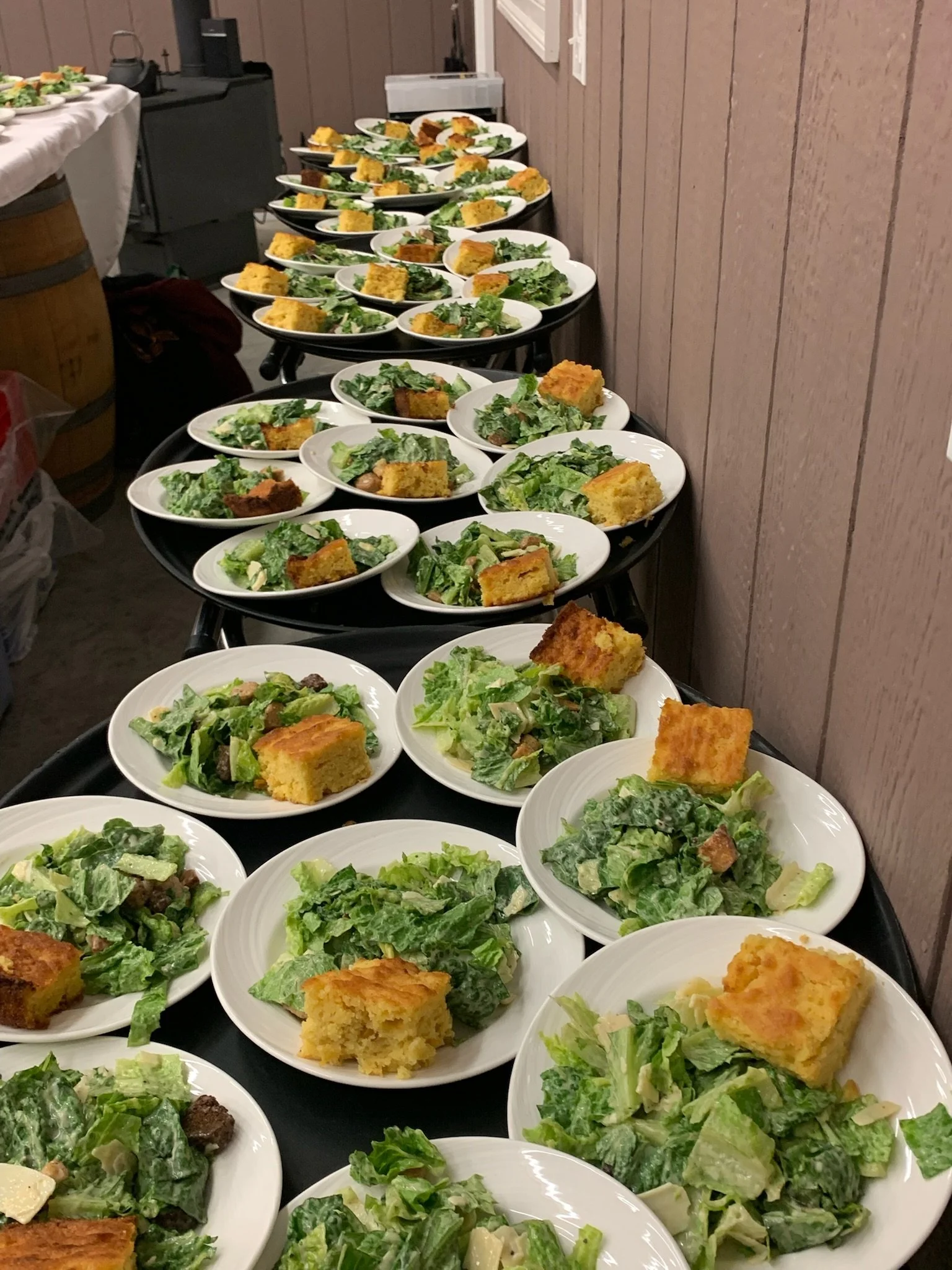 Multiple white plates with salad and slices of cornbread arranged on a long black table in a room with wooden walls.