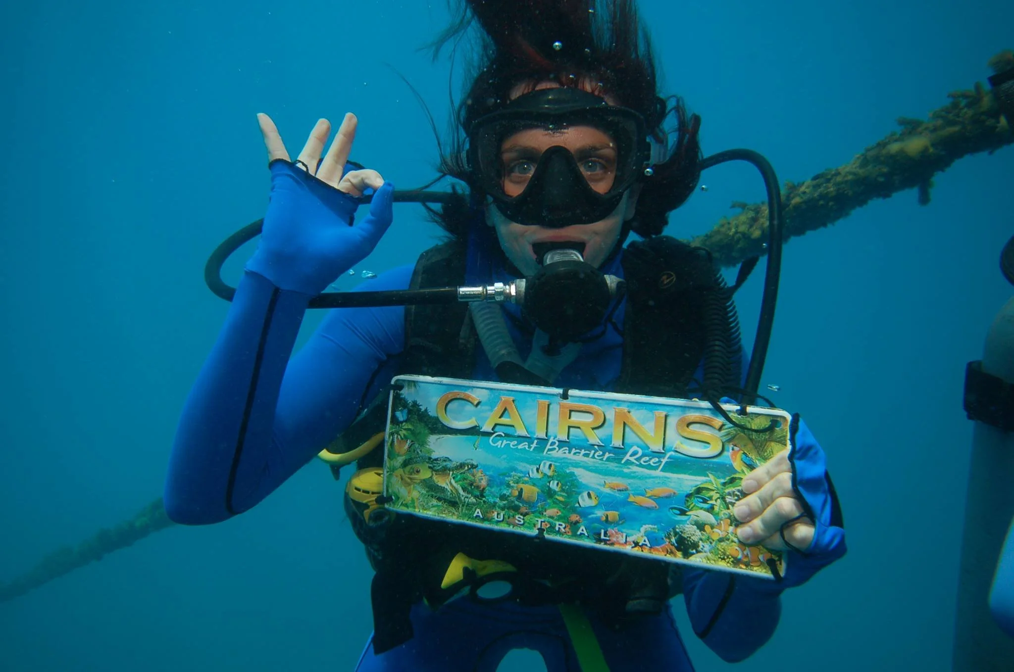A scuba diver underwater holding a Cairns Great Barrier Reef souvenir sign and making an OK gesture.
