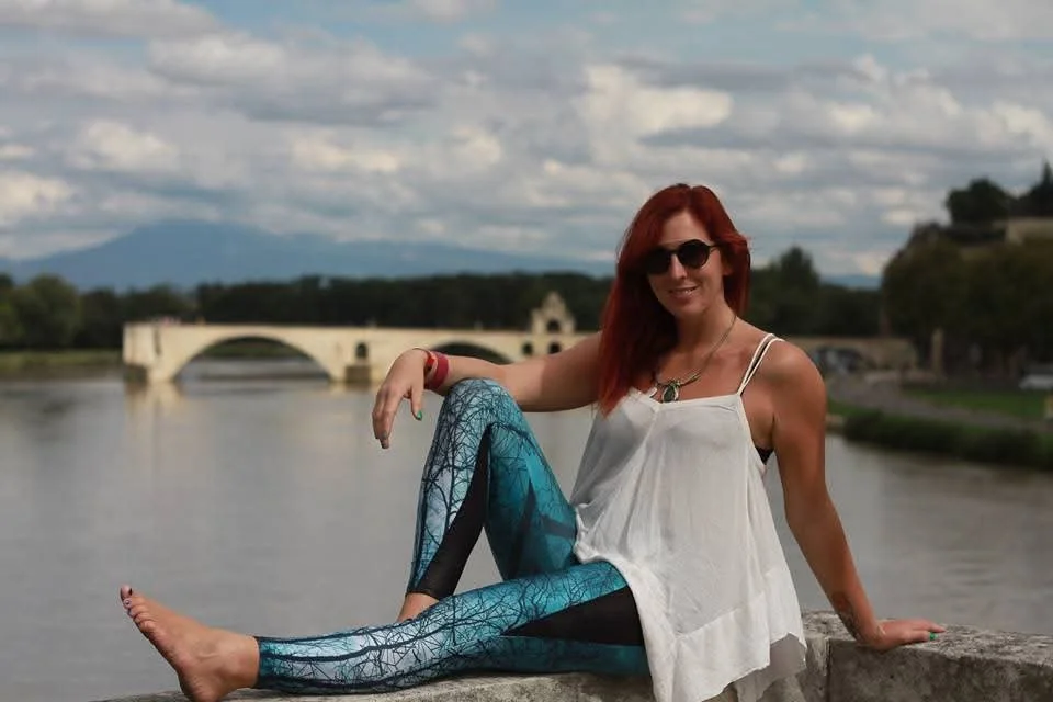 Woman with red hair wearing sunglasses, white top, and patterned leggings sitting on a stone ledge by a river with a bridge in the background.