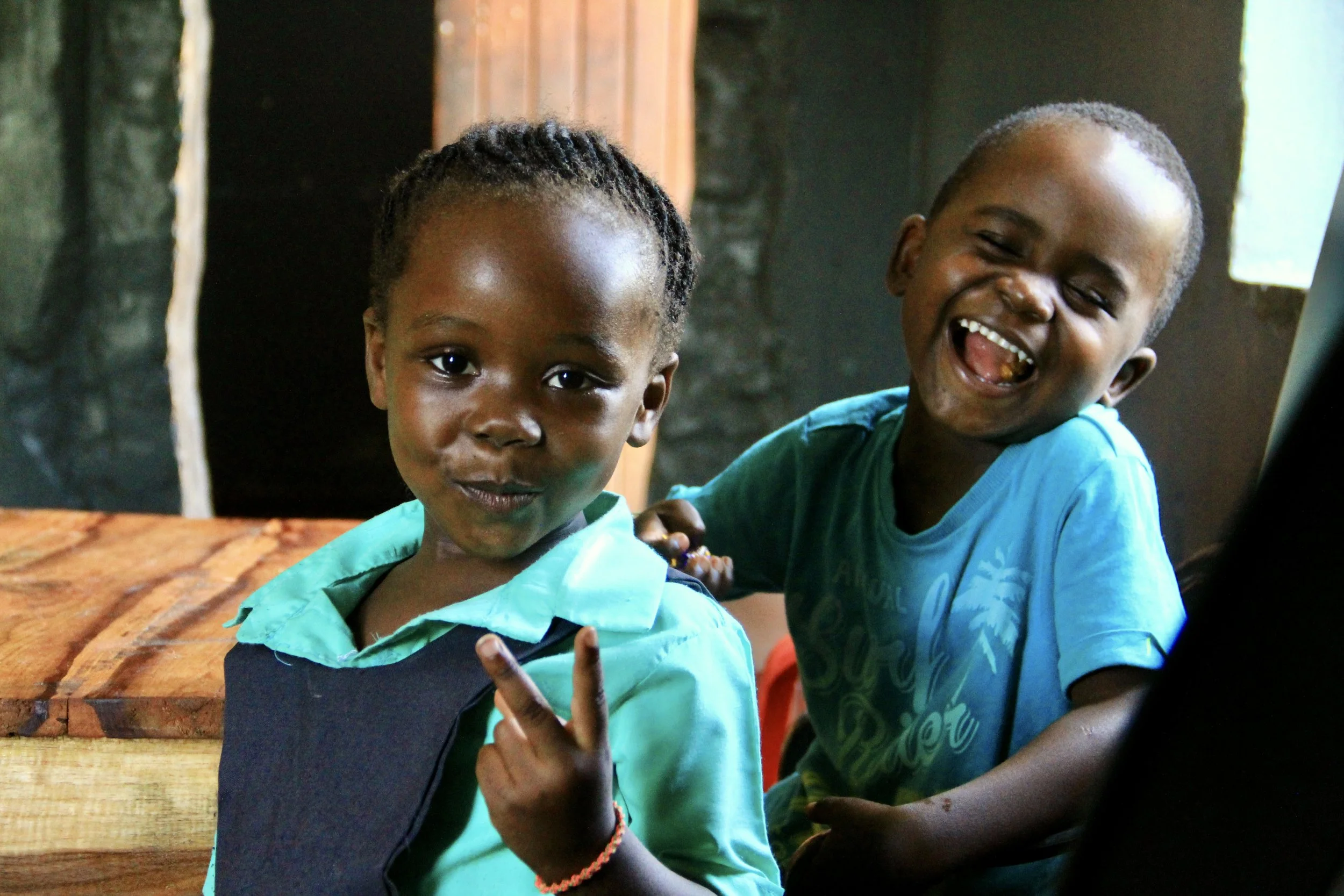 Two young children smiling and making peace signs, inside a room with dark walls and a window.