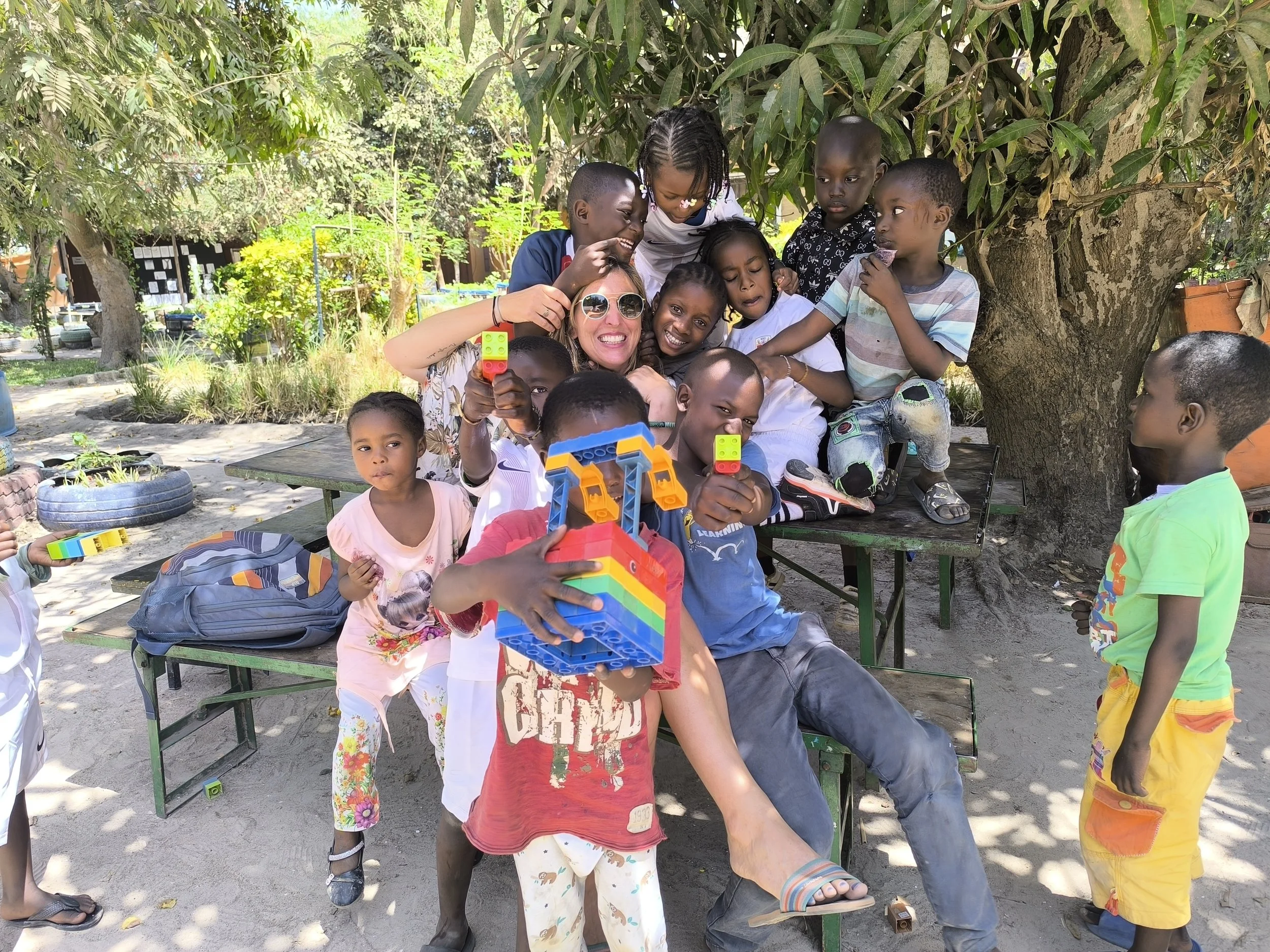 Group of children and a woman outdoors under a tree, playing with building blocks, holding up small colorful blocks, some sitting, some standing, and smiling.