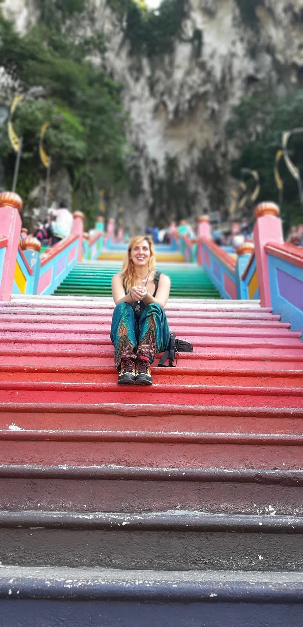 A woman sitting on colorful stairs leading up to a temple or cultural site, with a rocky cliff and greenery in the background, smiling and holding her hands together.