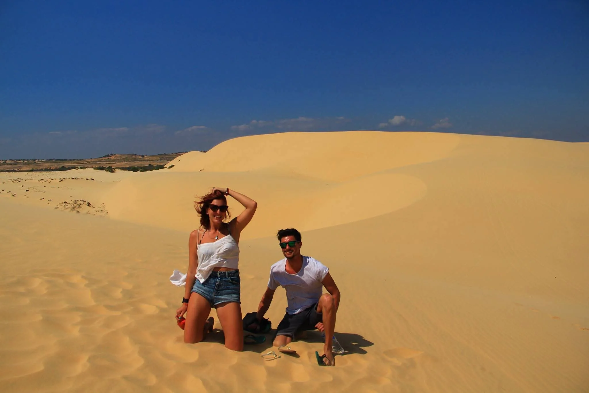Two young adults, a woman and a man, are posing on sandy desert dunes under a clear blue sky. The woman is standing with one hand in her hair wearing sunglasses, a white tank top, and denim shorts. The man is kneeling with one knee on the sand, weari