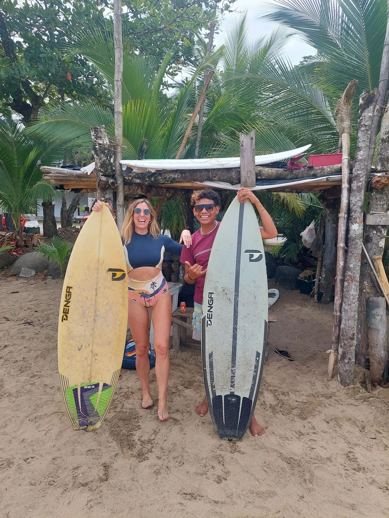 Two people on a sandy beach holding surfboards, surrounded by tropical palm trees and greenery, with a makeshift shelter behind them.
