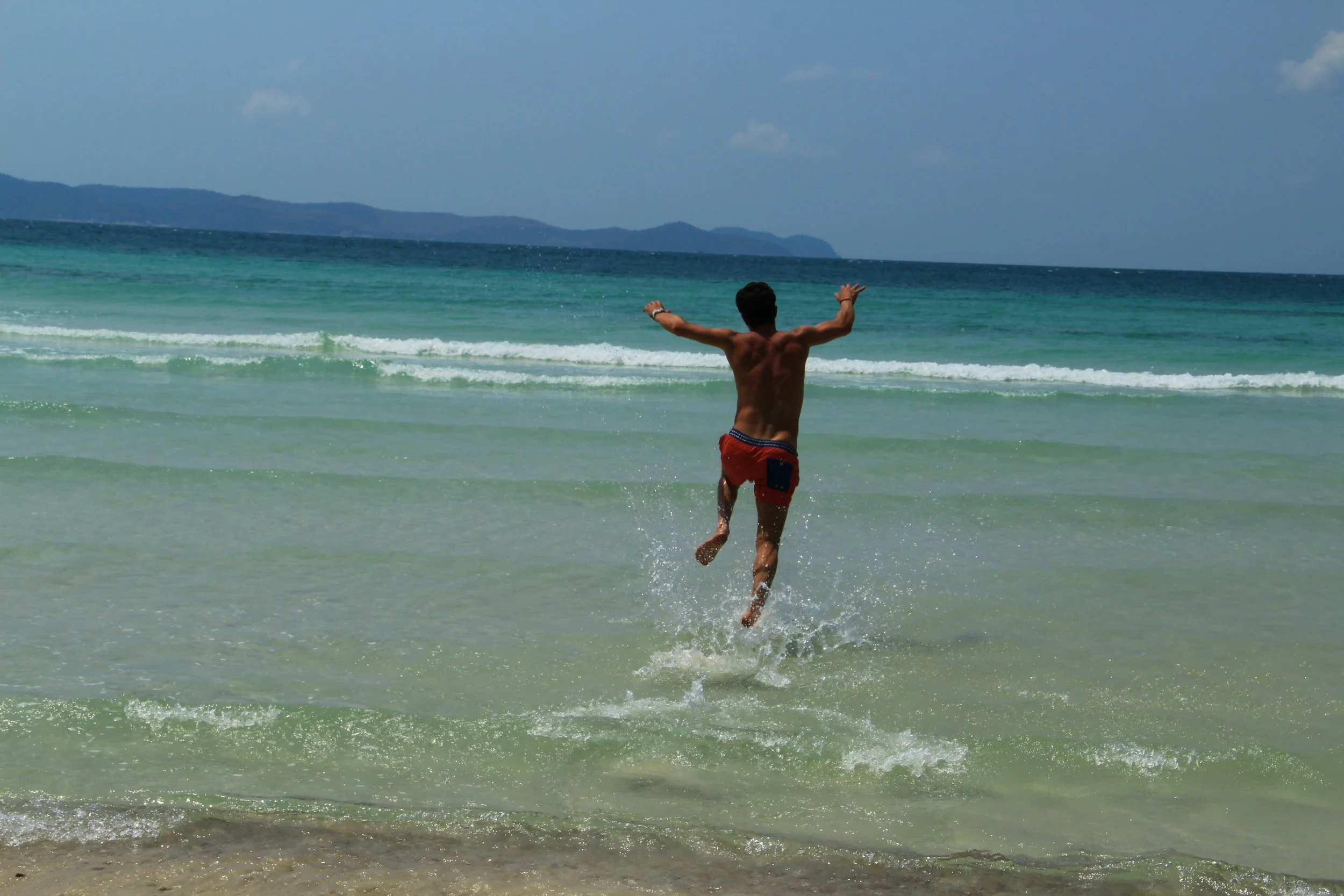 A person running into the ocean on a beach with arms outstretched and water splashing around.
