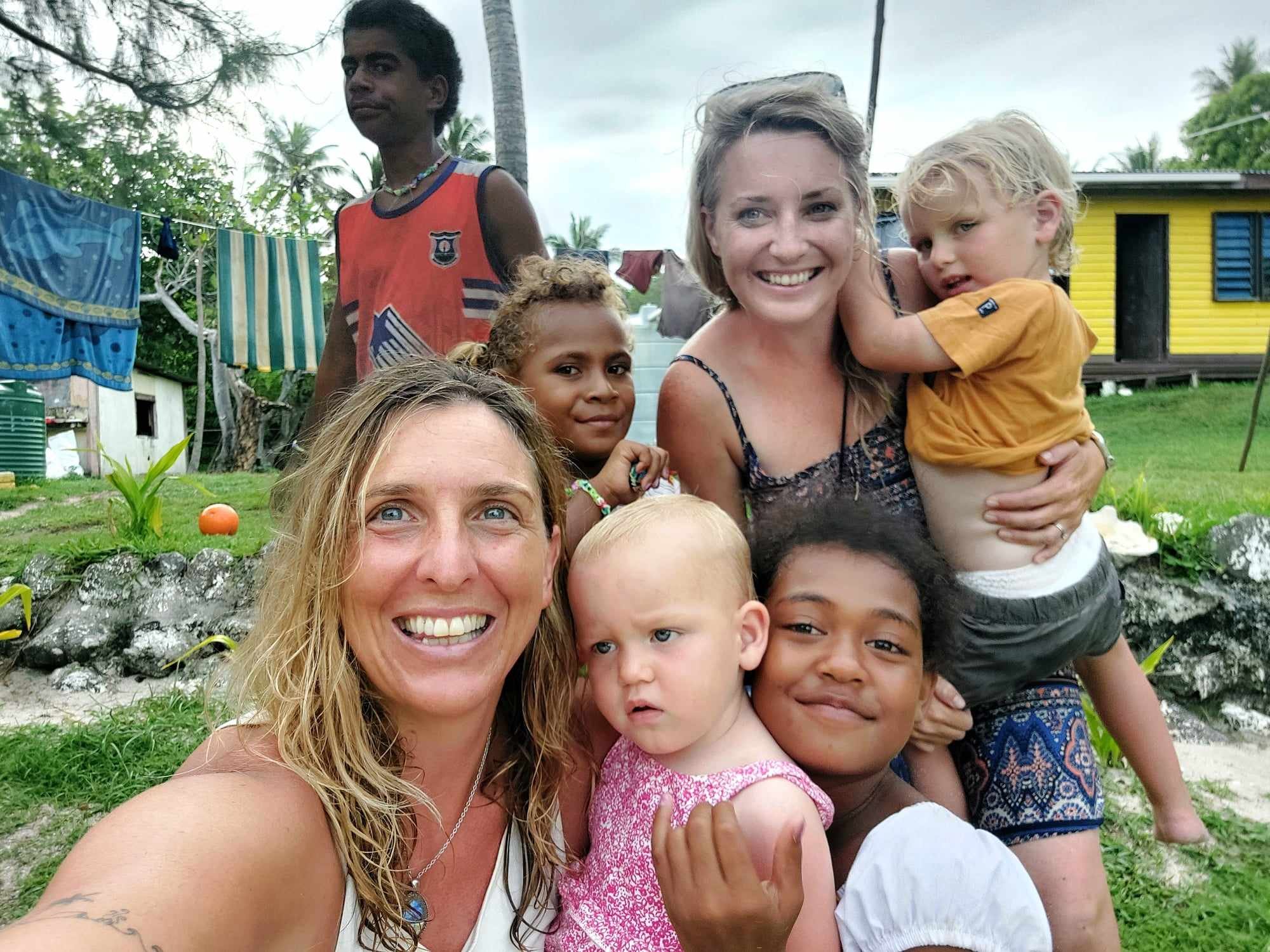 A group of smiling children and women posing outdoors in a tropical setting. There are three women and five children, with a colorful yellow house and laundry hanging on a line in the background.