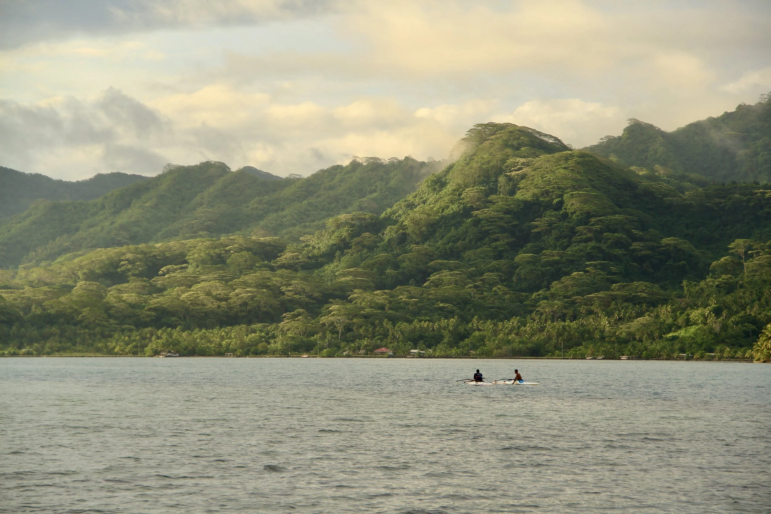 Two people paddleboarding on a large body of water with lush green mountains in the background and a partially cloudy sky.