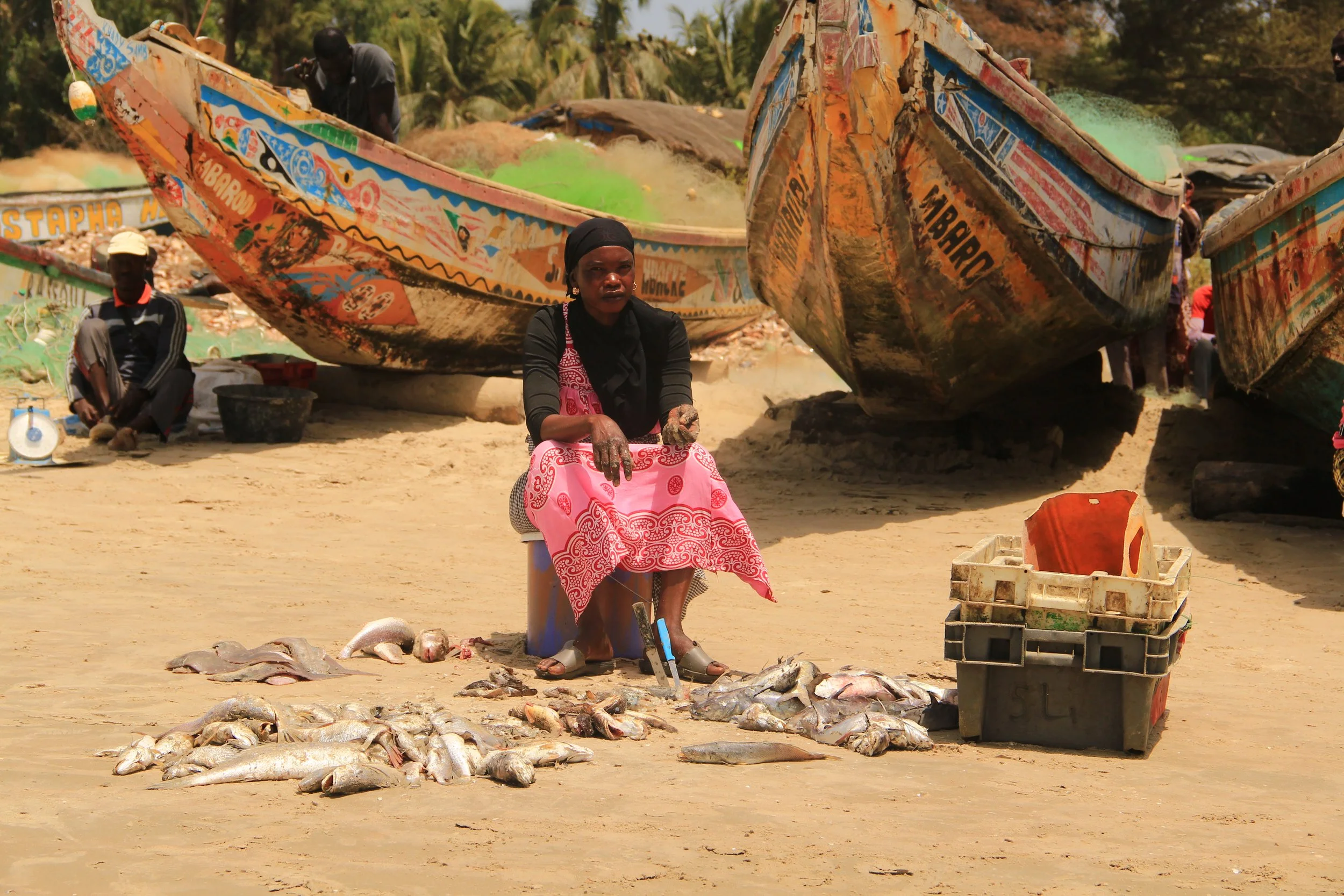 Woman selling freshly caught fish on the beach with boats in the background.