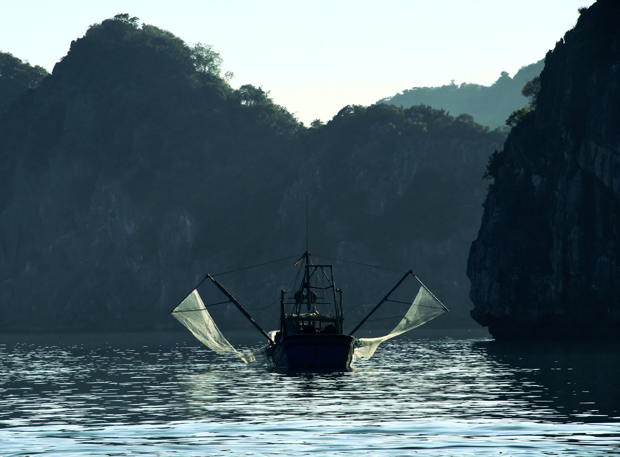 A fishing boat with large nets extends from the sides, floating on calm water surrounded by high rocky cliffs and lush green hills.