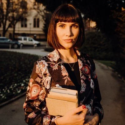 Young woman with short dark hair and bangs holding a stack of books outdoors during sunset.