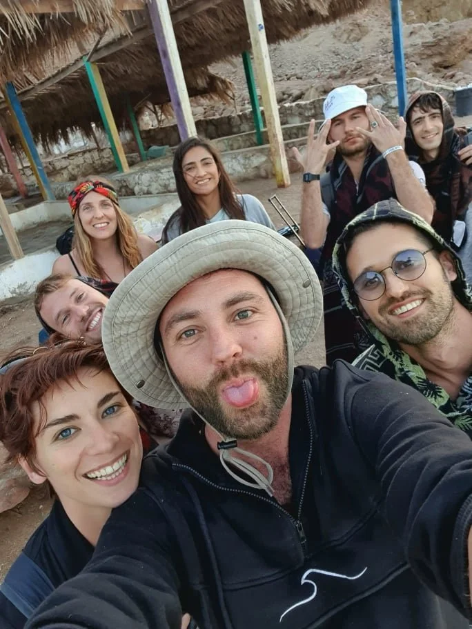 A group of seven friends taking a selfie outdoors at a beach with rustic structures and rocky terrain in the background. They are smiling, and some are making playful gestures.