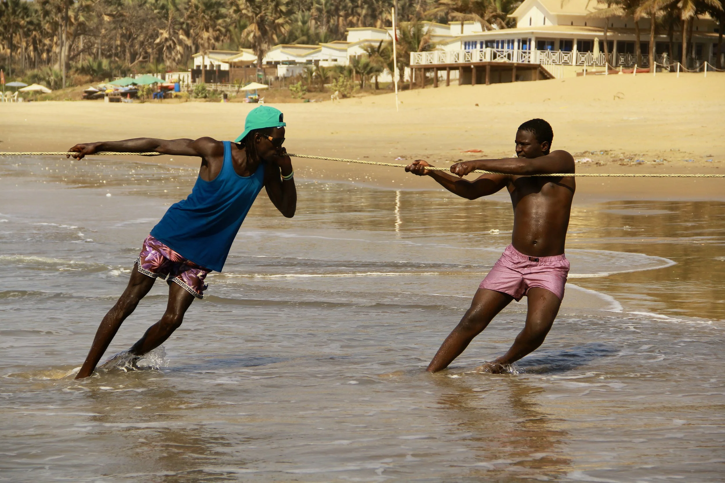 Two men are playing tug-of-war with a rope in shallow water on a beach, with buildings and palm trees in the background.