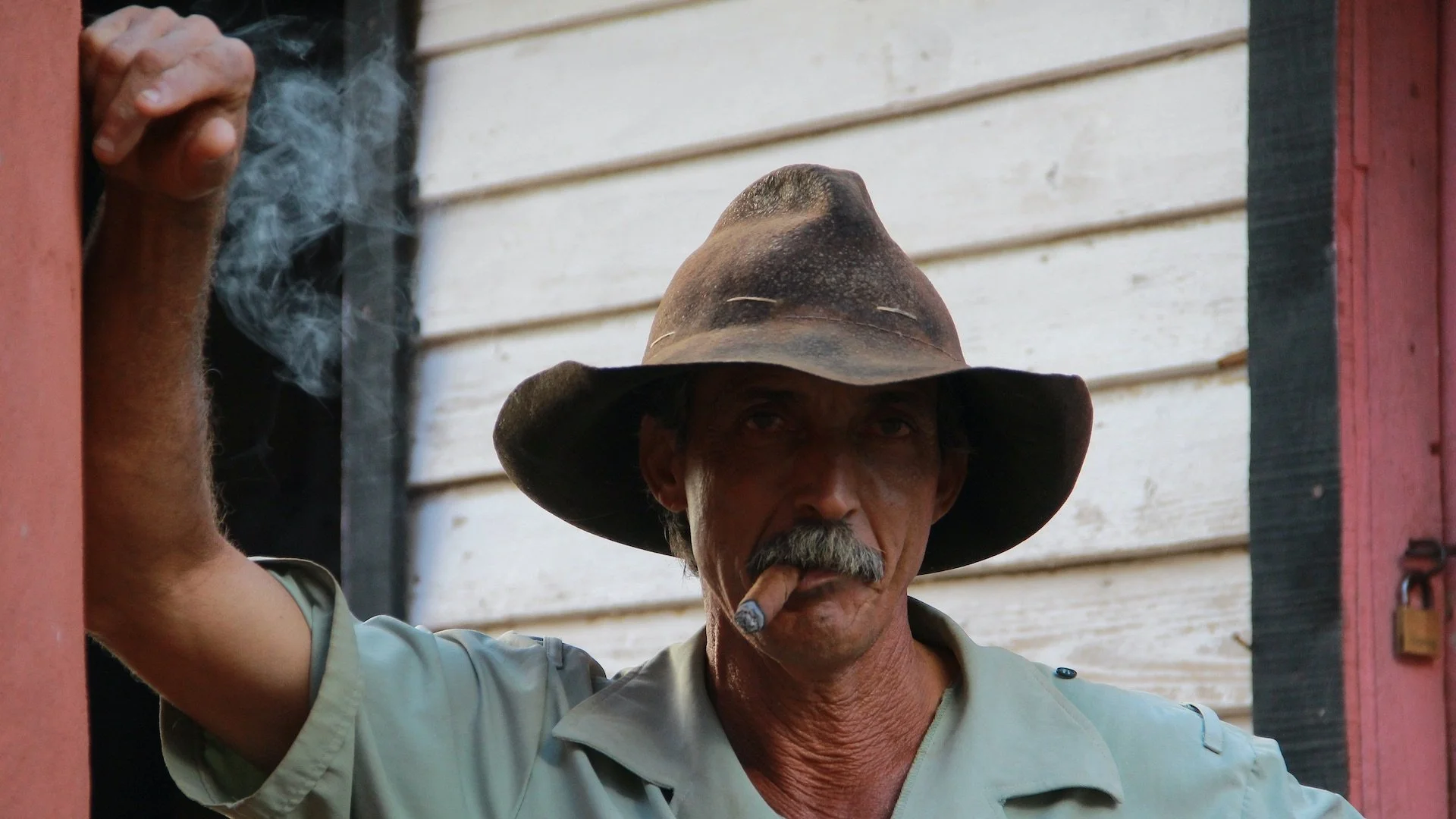 An older man with a mustache wearing a large, weathered cowboy hat and smoking a cigar. He is standing outside, leaning on a doorframe, with a background of white painted wooden siding.