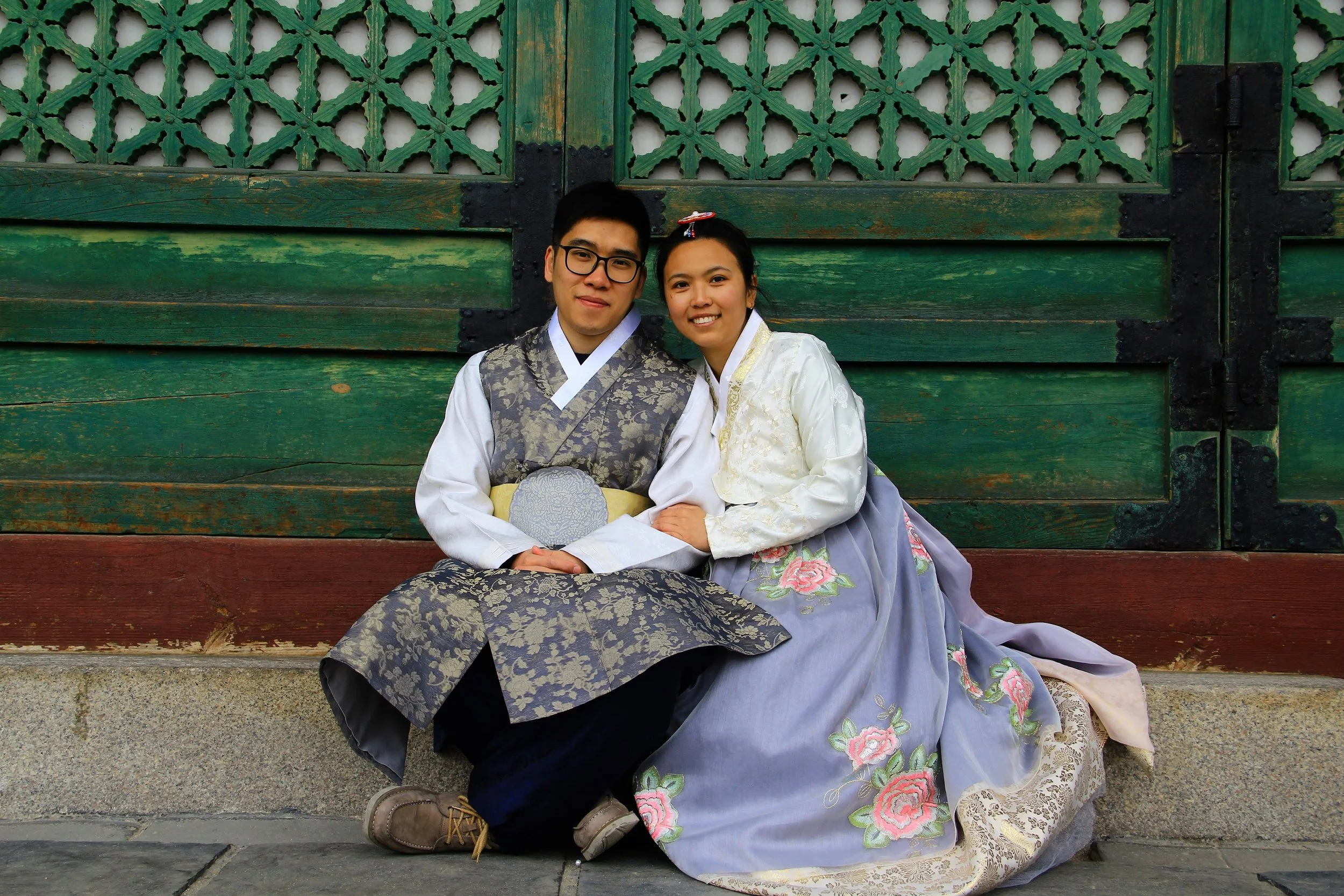 A young couple dressed in traditional Korean hanbok sitting beside a green wooden gate with decorative patterns.