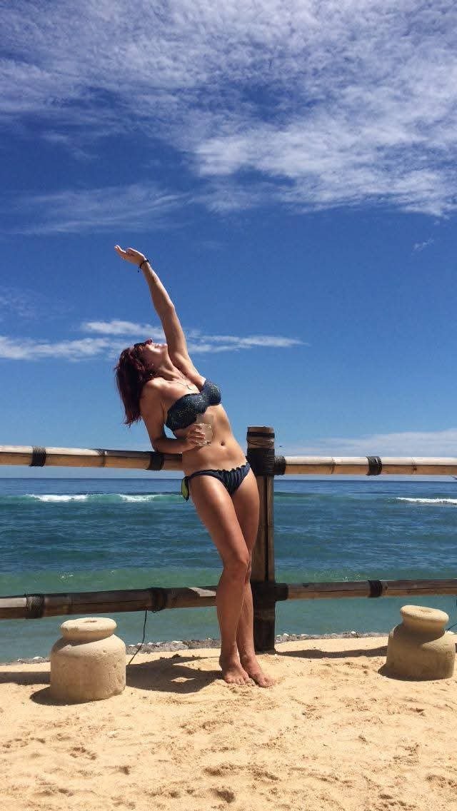Woman in black bikini standing on sandy beach, leaning against wooden railing, stretching arm upward, with ocean and sky in background.