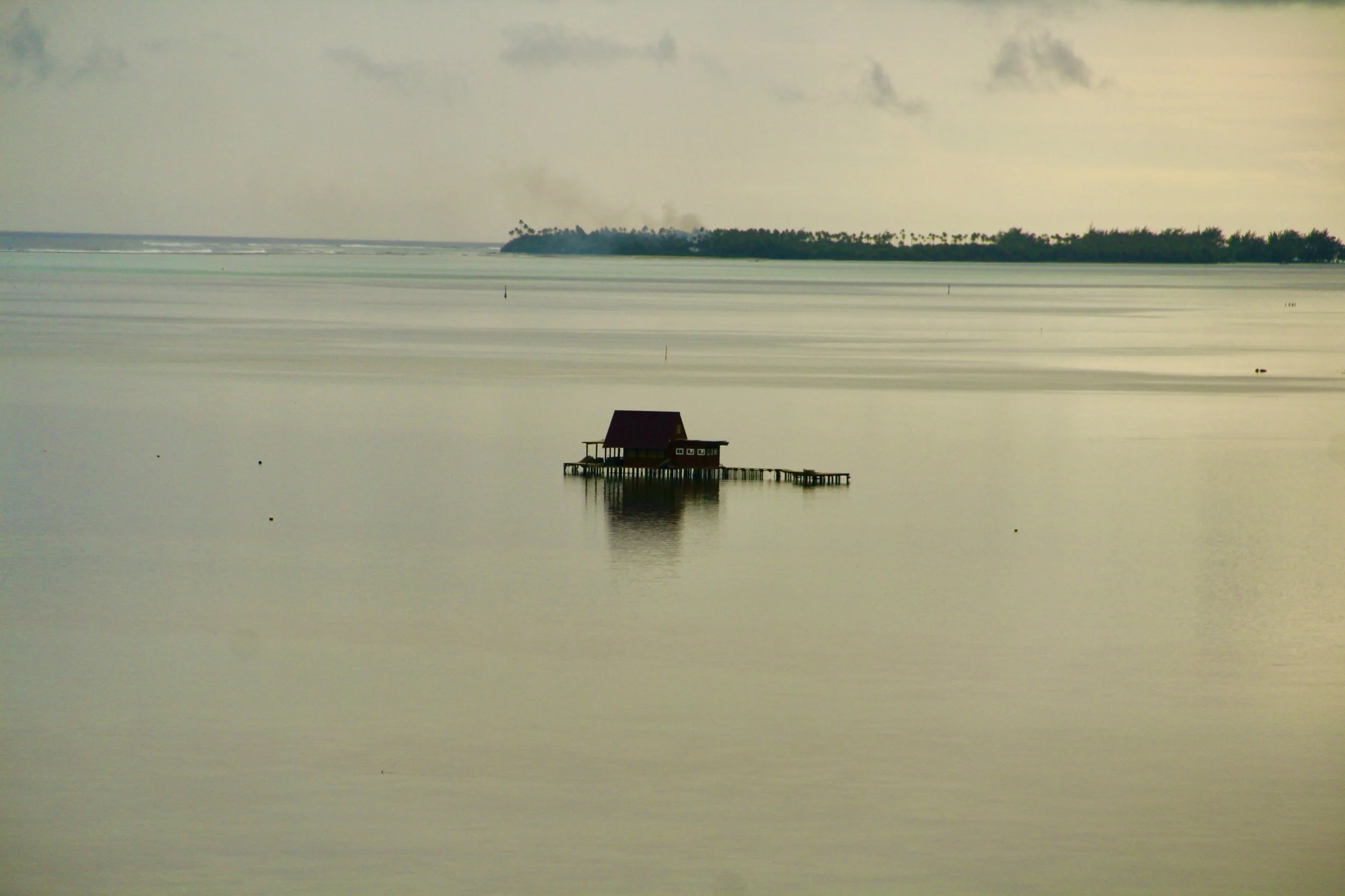 A house on stilts over calm water with an island in the background and a cloudy sky.